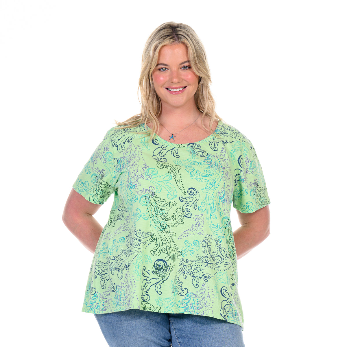 A woman with blonde hair wearing the Waverley Swirl Luna Top—a light green, flowy short-sleeve tee crafted from soft cotton slub by Fresh Produce Clothes in plus size—paired with blue jeans smiles at the camera with her hands behind her back against a white background.