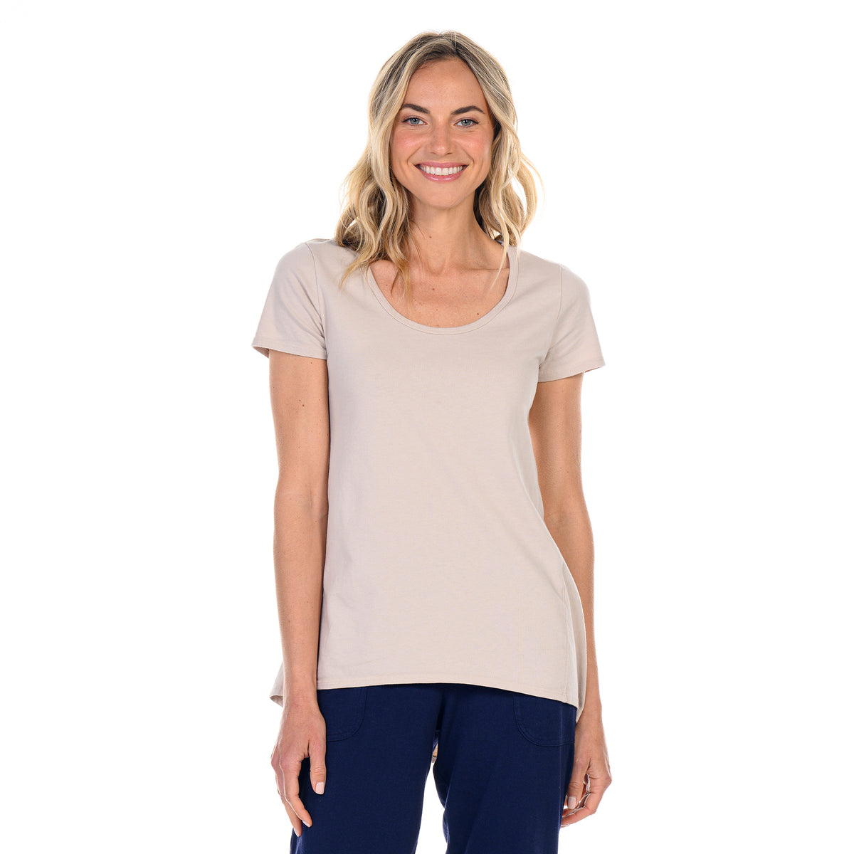 A woman with long, wavy blonde hair wearing the Luna Top by Fresh Produce Clothes—a flowy, short-sleeve tee crafted from mid-weight cotton slub—paired with dark blue pants smiles while standing against a white background.