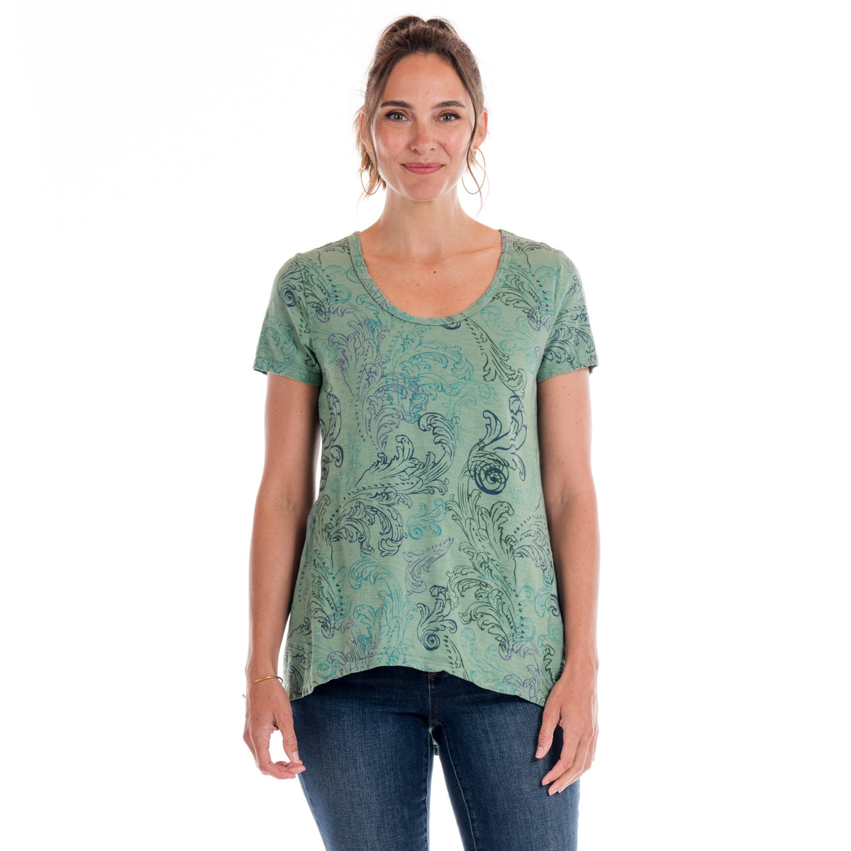 A woman stands facing forward, wearing the Fresh Produce Clothes Waverley swirl Luna Top in plus size—a green, flowy short sleeve tee crafted from patterned cotton slub fabric—paired with blue jeans. Her light brown hair is tied back and she’s smiling gently against a white background.