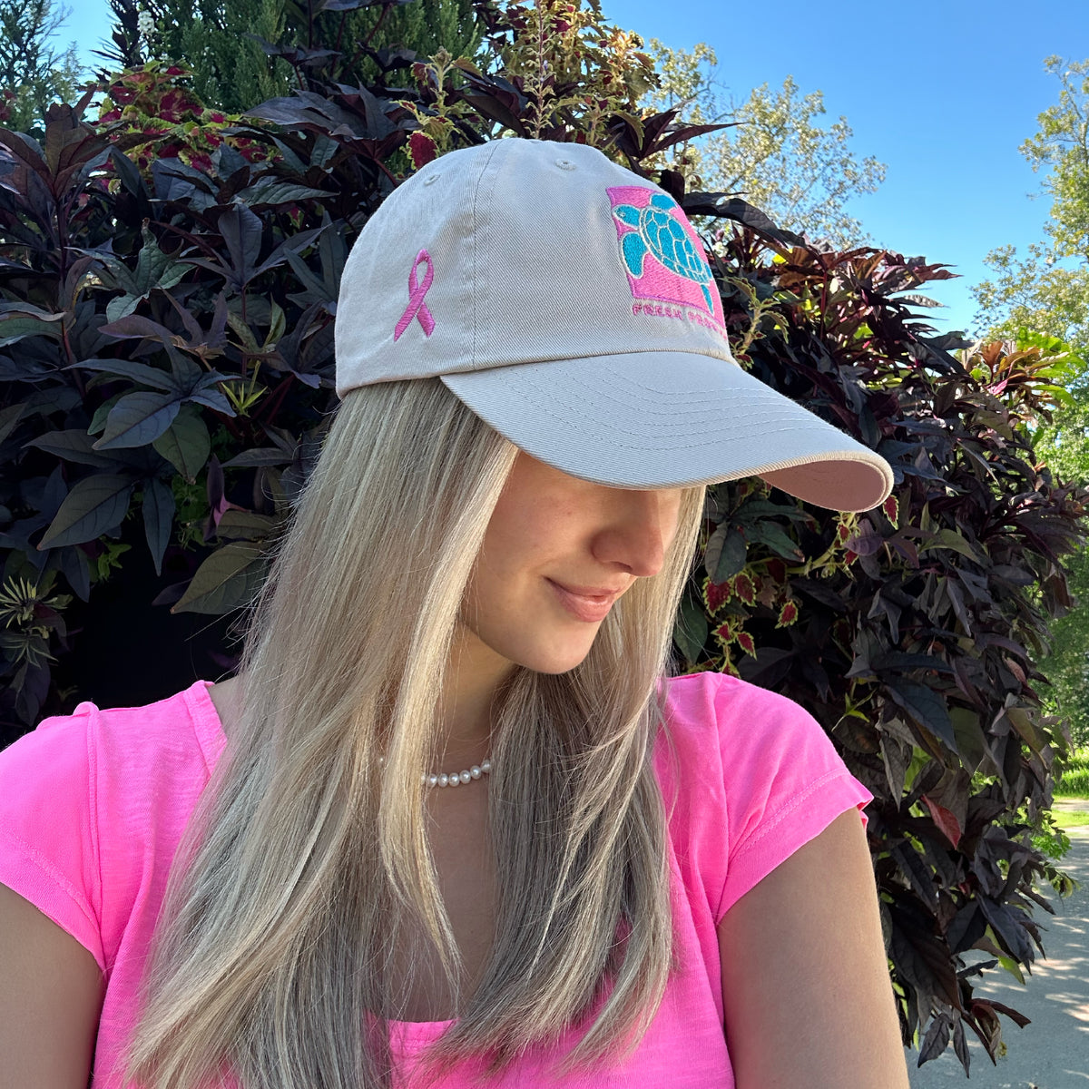 A woman with long blonde hair wears the Breast Cancer Turtle Hat by Fresh Produce Clothes and a pink shirt, standing outdoors near dark-leaved plants, looking down with a slight smile.