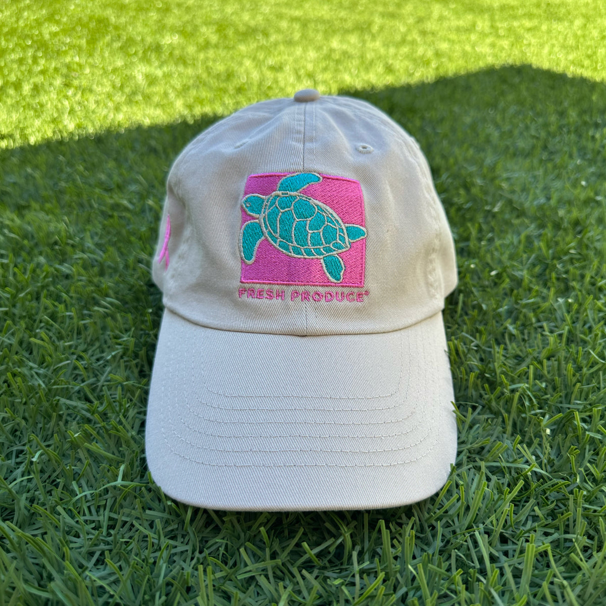 The Breast Cancer Turtle Hat by Fresh Produce Clothes, featuring a turquoise turtle and brand embroidery, is shown on green artificial grass.