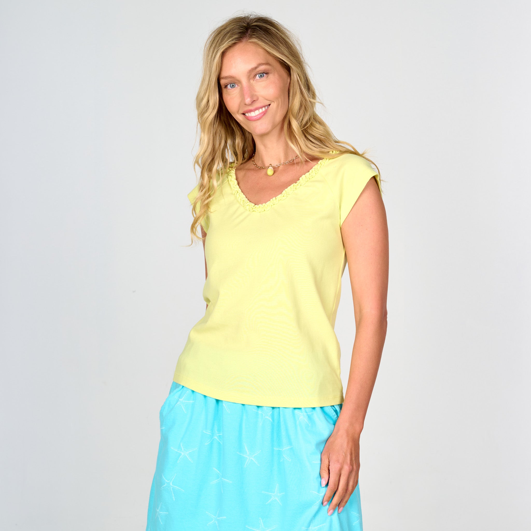 A woman with long blonde hair is smiling while wearing the Fresh Produce Clothes Baby Rib Ruffle V Neck T in yellow, a cap-sleeve top crafted from soft baby rib fabric, paired with a light blue skirt featuring white star patterns, standing against a plain light gray background.
