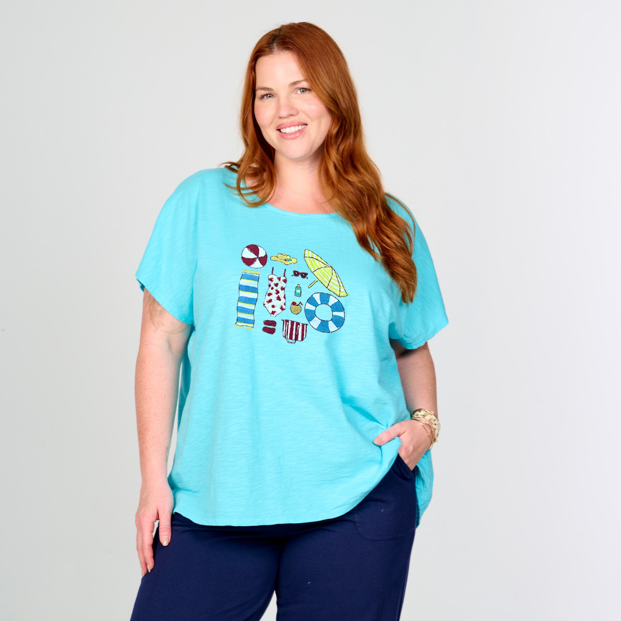 A woman with long red hair smiles with one hand in her pocket, modeling the Fresh Produce Clothes Beach Day Essentials Keepsake T | Plus Size—featuring colorful beach-themed graphics on light blue and paired with dark blue pants—against a plain light background for an effortless summer vibe.