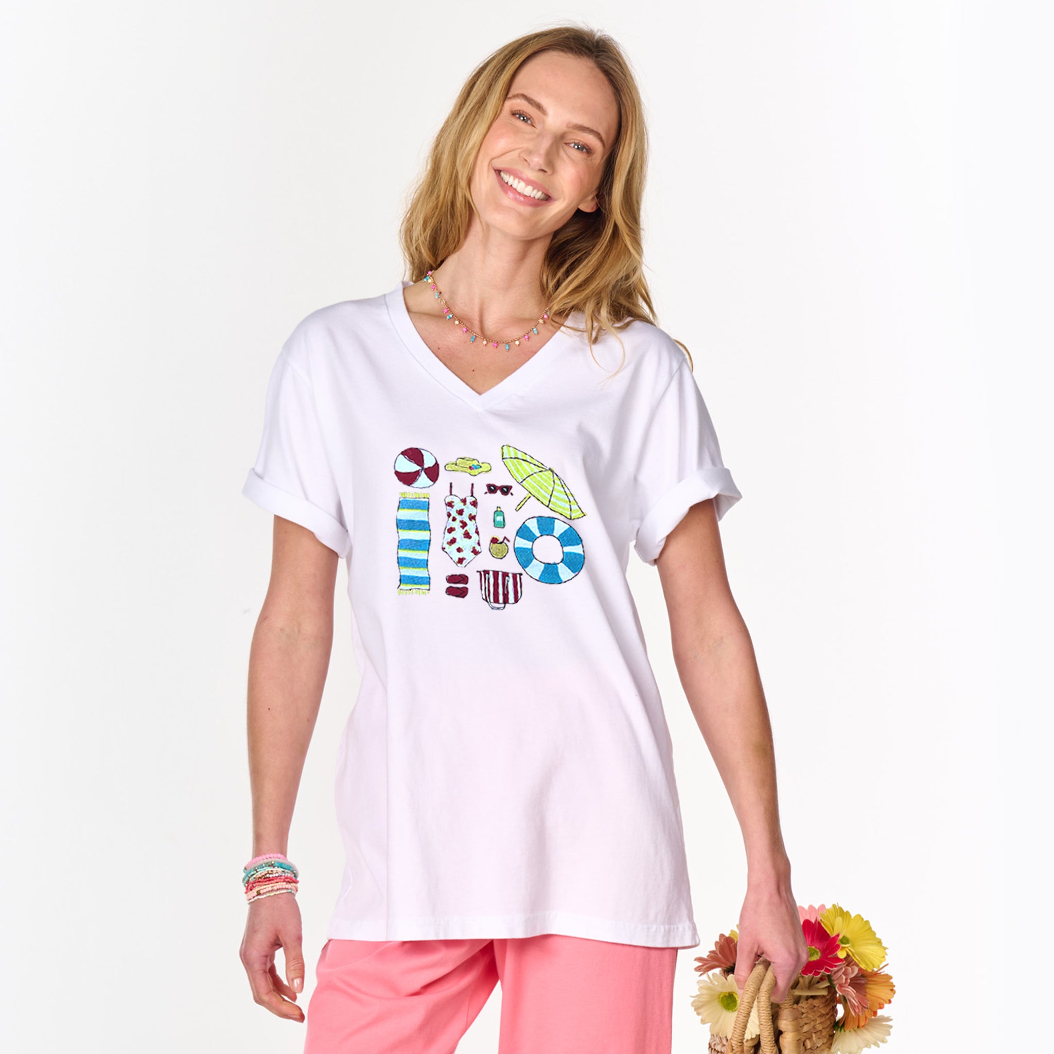A smiling woman wears the Fresh Produce Clothes Beach Day Essentials V-Neck T in white, featuring colorful beach-themed graphics. She pairs it with pink pants and carries a straw bag decorated with yellow and pink flowers.