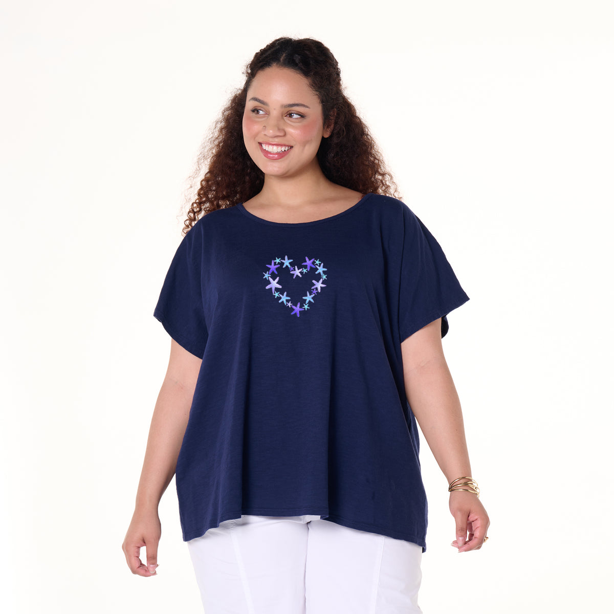 A smiling woman with curly hair models the Blue Sea Heart Keepsake T | Plus Size by Fresh Produce Clothes, showcasing its loose navy blue design and starry heart graphic. Paired with white pants and set against a plain white background, this tee makes the perfect beach gift for someone special.