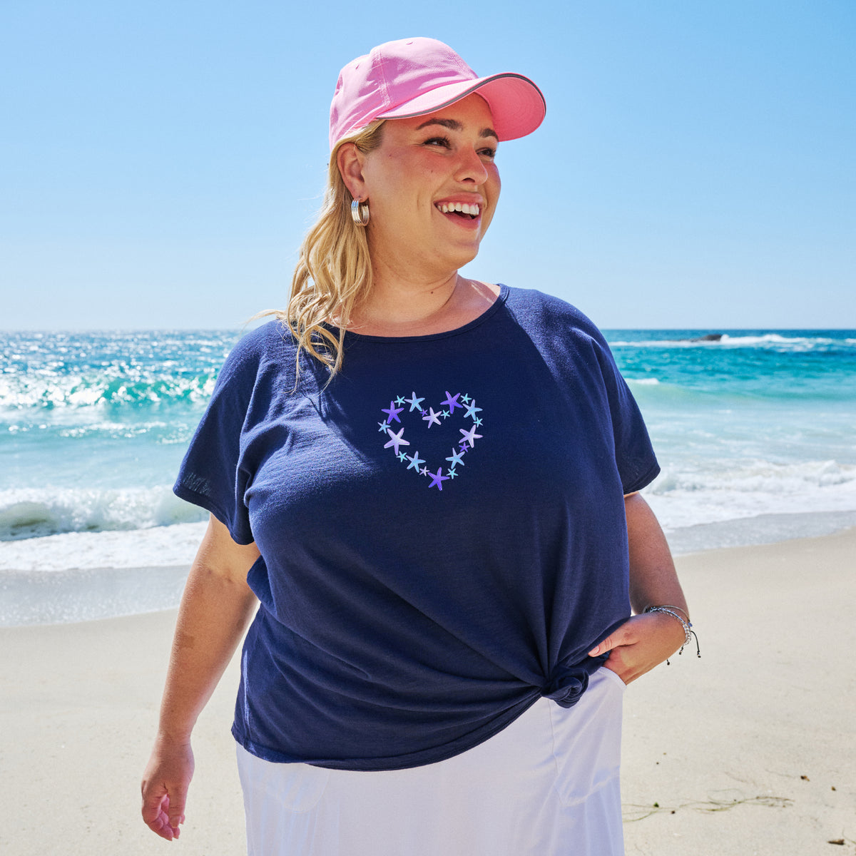 A woman in a pink cap and navy Blue Sea Heart Keepsake T | Plus Size from Fresh Produce Clothes stands smiling on a sunny beach, with waves and blue sky in the background—a perfect scene for a thoughtful beach gift.