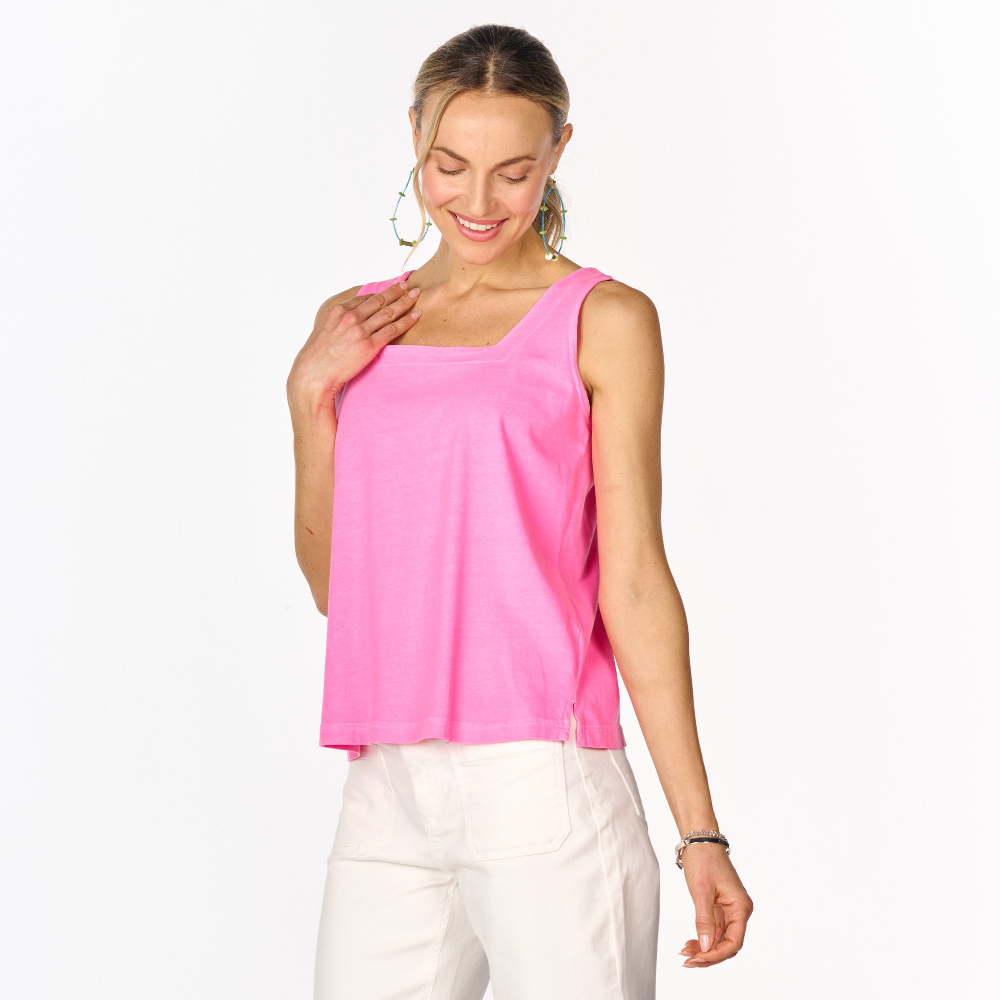 A woman wearing the Boxy Tank by Fresh Produce Clothes in a bright pink hue, paired with white pants, smiles while looking down and stands against a plain white background.