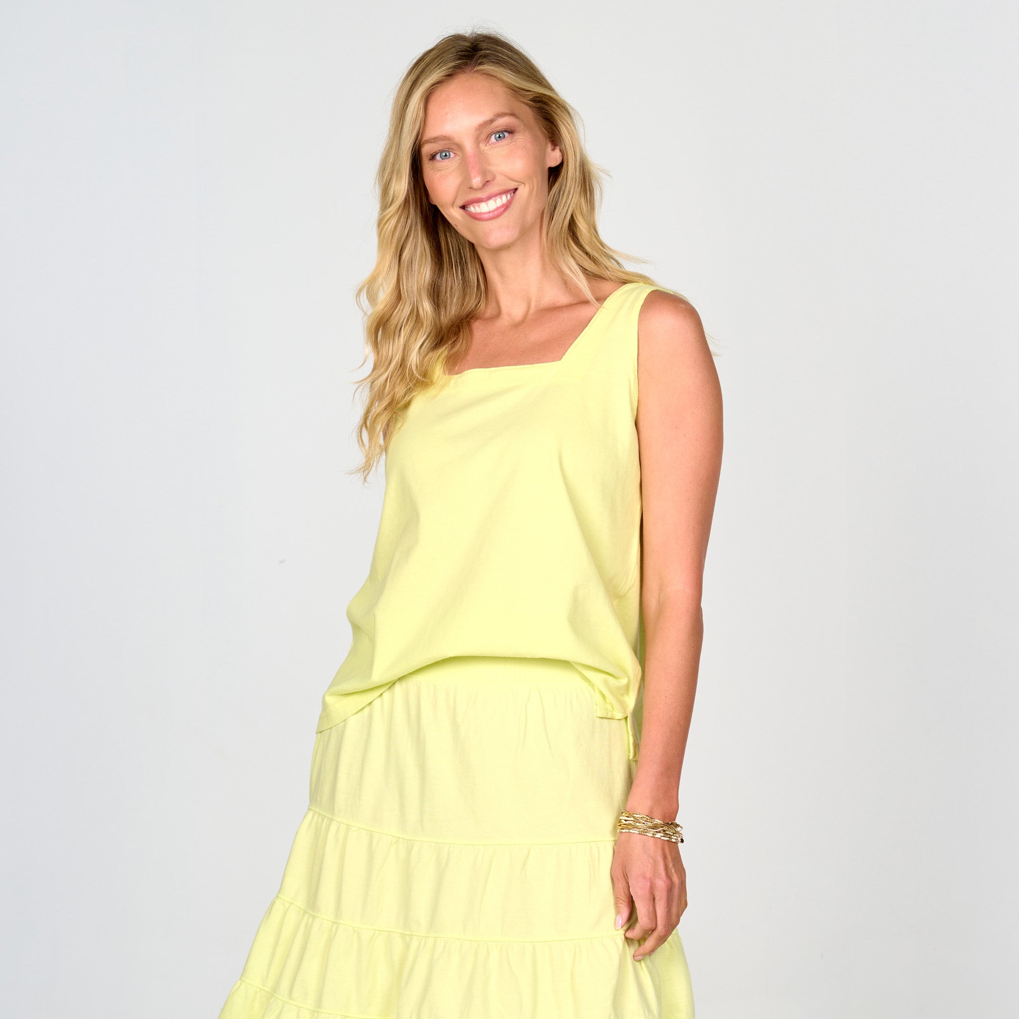 A woman with long blonde hair is smiling while standing against a plain light-colored background, wearing the Fresh Produce Clothes Boxy Tank in a bra-friendly, sleeveless yellow square neck style, paired with a matching yellow tiered skirt.