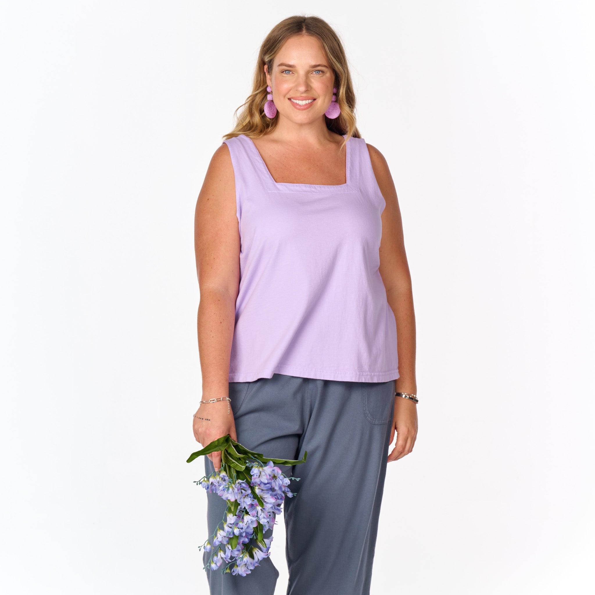 A woman wearing the Fresh Produce Clothes Boxy Tank in lavender—a bra-friendly, sleeveless top with a relaxed silhouette—pairs it with gray pants and stands smiling against a plain white background while holding a bunch of purple flowers. This plus size tank, now on clearance, offers effortless style and comfort.