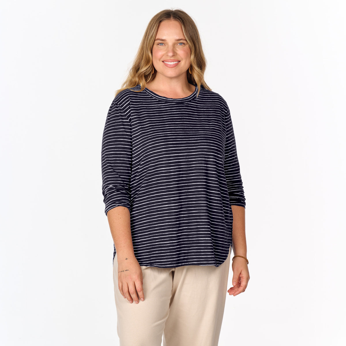 A smiling person with long hair is wearing the Fresh Produce Clothes Ocean Stripe Catalina Top in plus size and beige pants, standing against a plain white background.