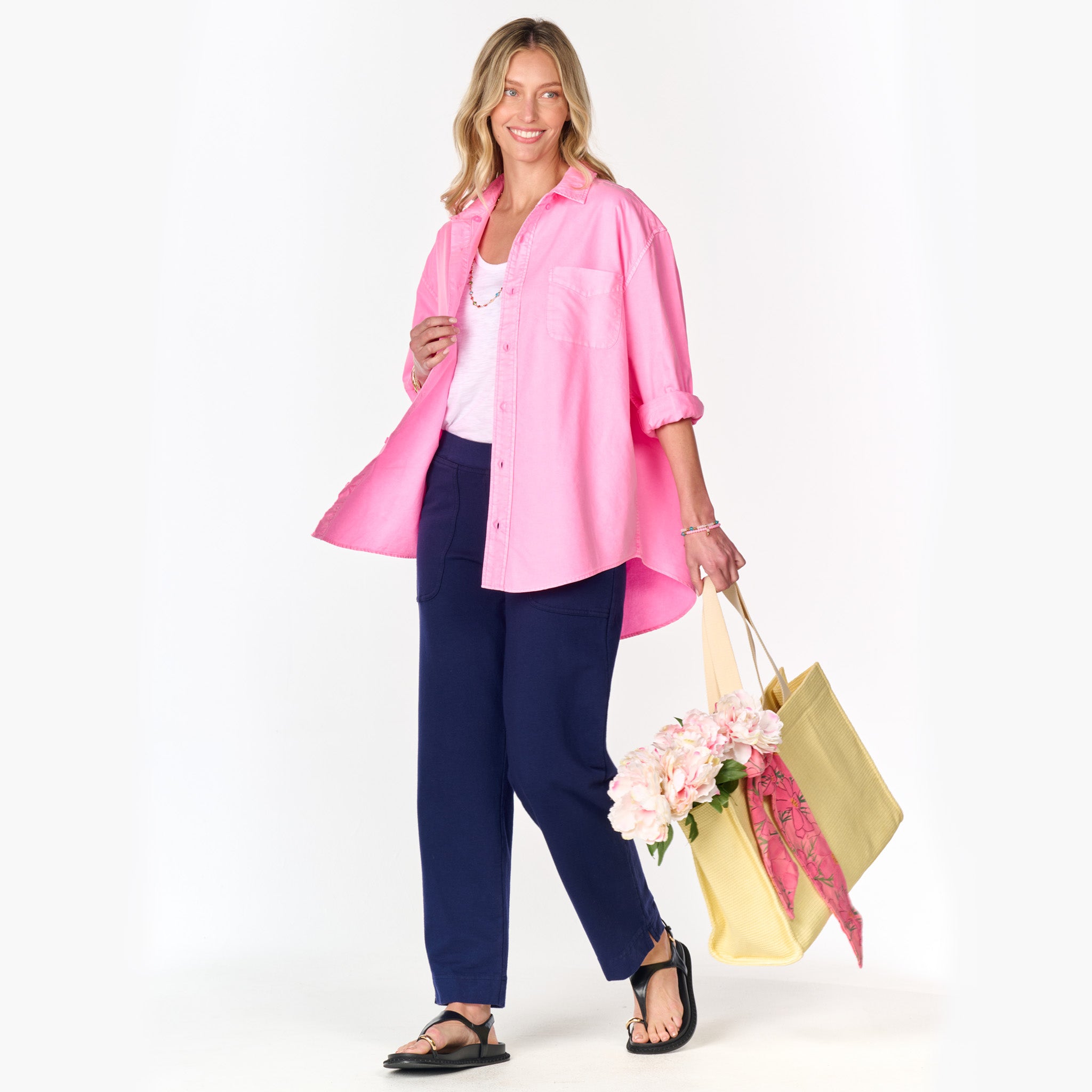 A smiling woman wears the Classic Oxford Shirt in bright pink from Fresh Produce Clothes (Clearance), styled oversized over a white top and navy pants. She holds yellow tote bags filled with flowers and a pink scarf, accessorized with black sandals and jewelry, standing against a white background.
