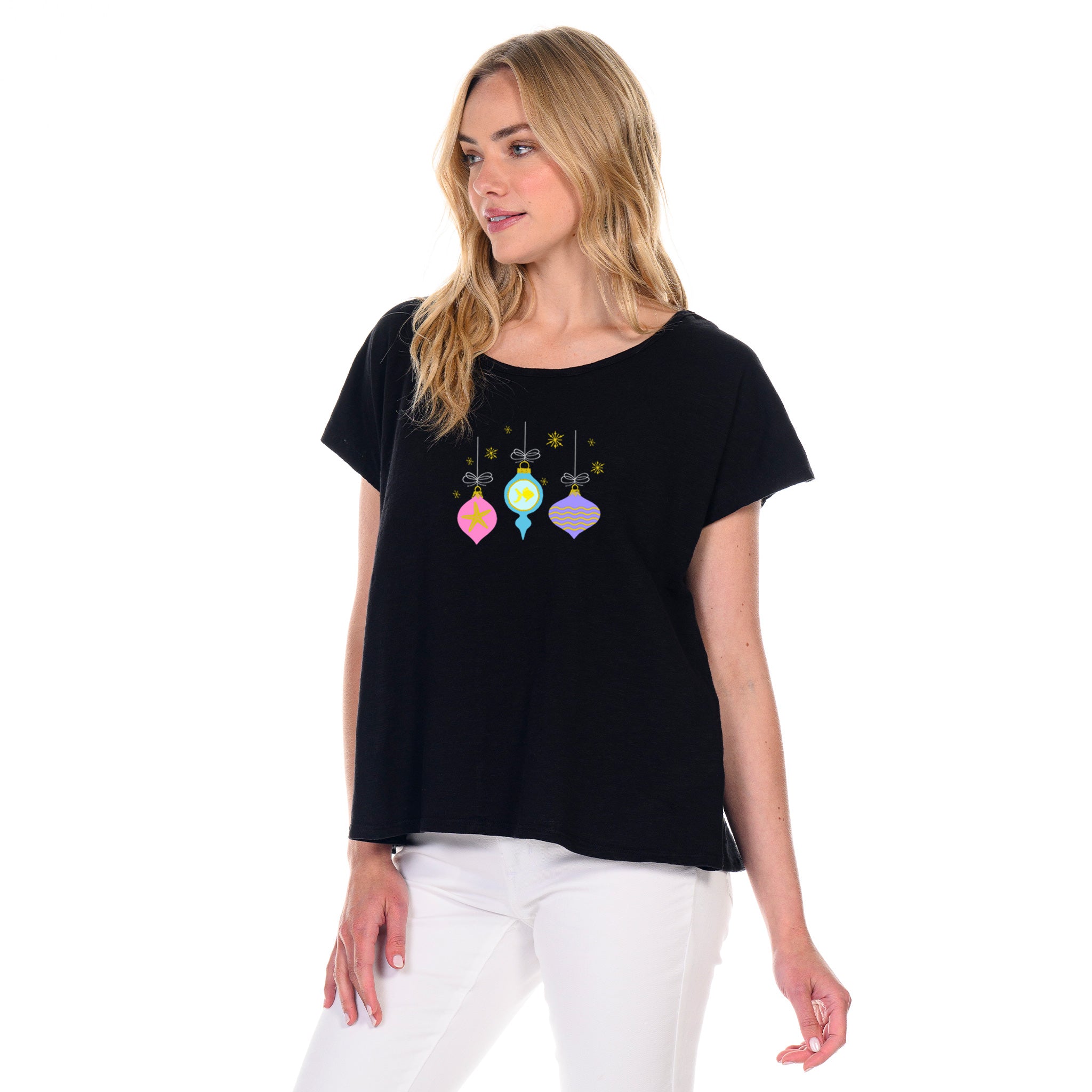 A woman stands against a white background, wearing the Fresh Produce Clothes Coastal Ornaments Keepsake T—its black fabric adorned with colorful ornament designs that capture a festive holiday spirit—and pairs it with crisp white pants.