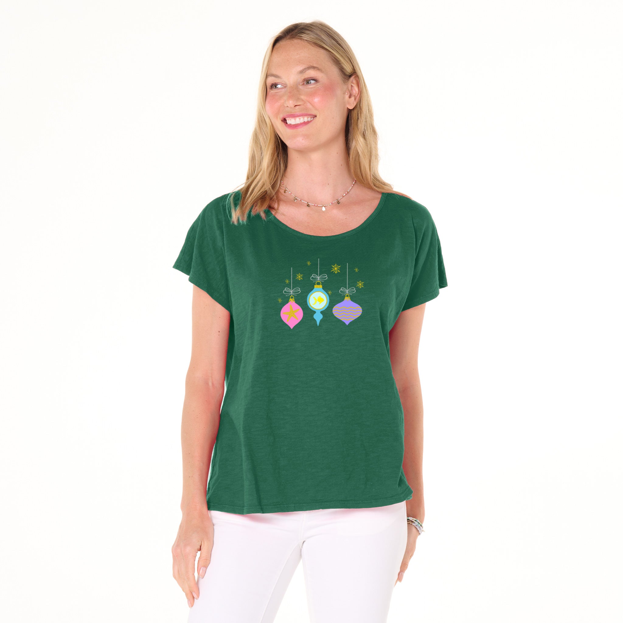 A smiling woman stands against a white background wearing white pants and the Fresh Produce Clothes Coastal Ornaments Keepsake T, featuring colorful hanging ornament designs—perfect for capturing the holiday spirit.