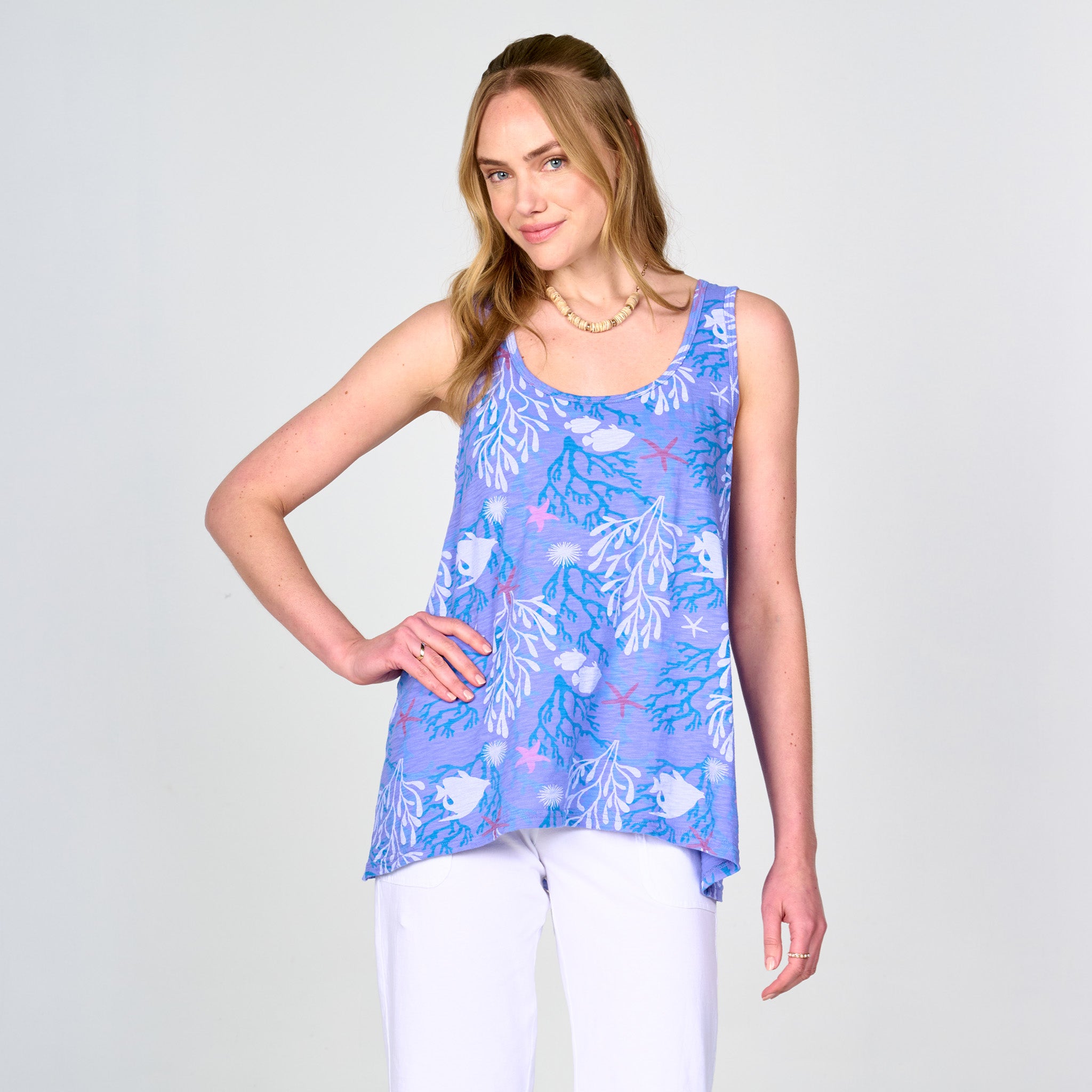 A woman stands against a plain light background, smiling with one hand on her hip, wearing the Fresh Produce Clothes Coral Reef Crossback Tank—a sporty blue patterned style—paired with white pants.