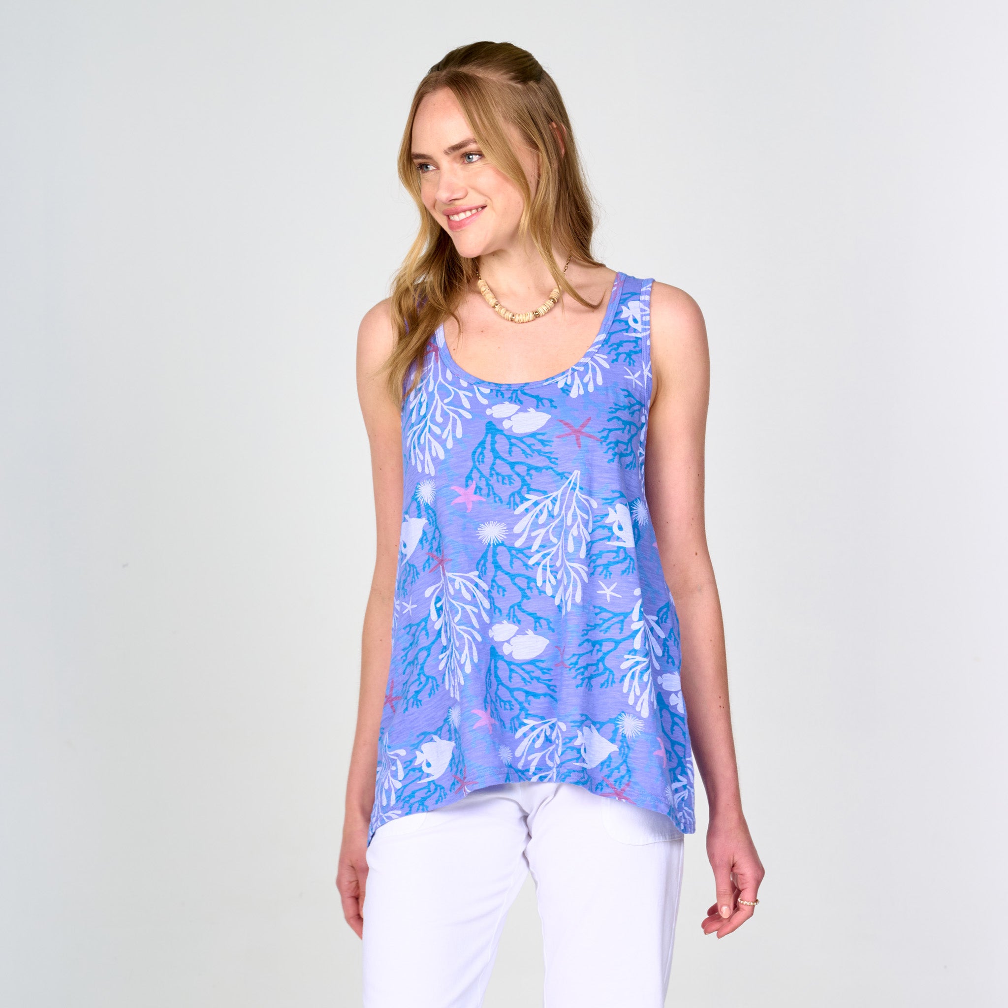 A woman wearing the Fresh Produce Clothes Coral Reef Crossback Tank Plus—a sleeveless sporty crossback tank featuring a blue and purple coral reef pattern and a fit-and-flare hem—paired with white pants, stands against a plain light background, smiling and facing slightly to the left.