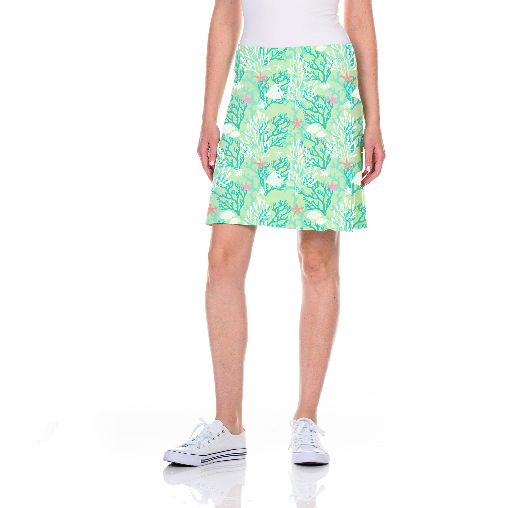 A person wearing a white top and the Coral Reef Marina Skirt by Fresh Produce Clothes—a mint julep green skirt with a botanical print, featuring an A-line silhouette and a flattering waistband—standing against a white background and wearing white sneakers.