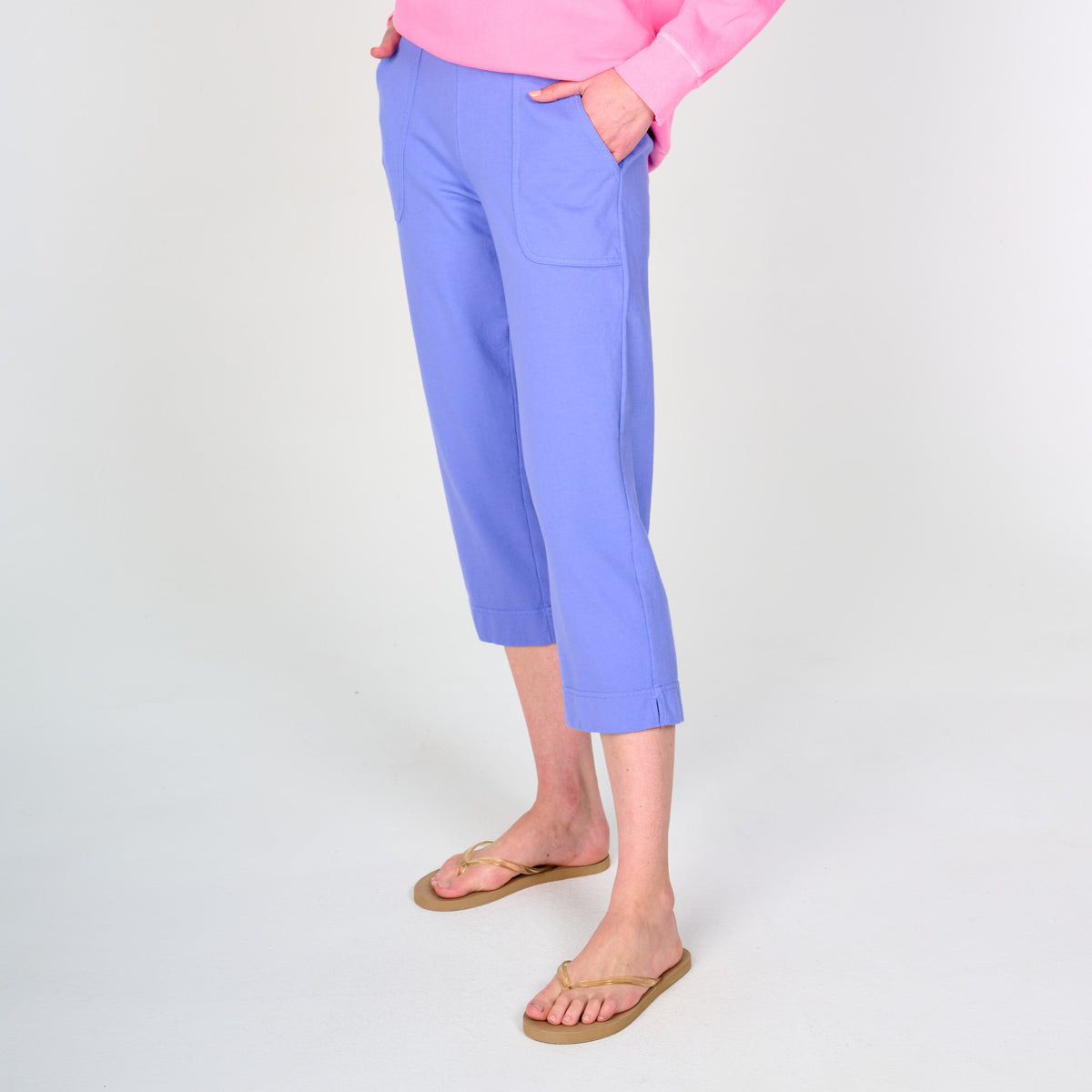 A person is shown from the waist down, standing against a plain white background and showcasing a relaxed, resort-ready style. They are wearing light purple Fresh Produce Clothes Coronado Key Largo Capris with pockets, paired with a pink long-sleeve top and beige flip-flops.
