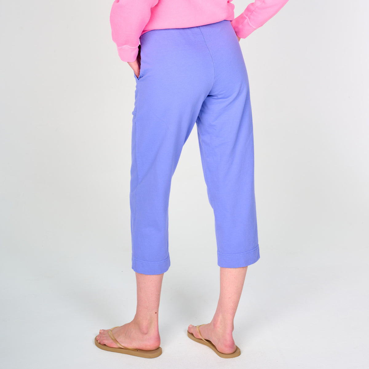 Wearing the Fresh Produce Clothes Coronado Key Largo Capris in light purple, paired with a pink long-sleeve top and tan sandals, this individual stands with hands in pockets facing away from the camera against a plain white background—perfectly resort ready.