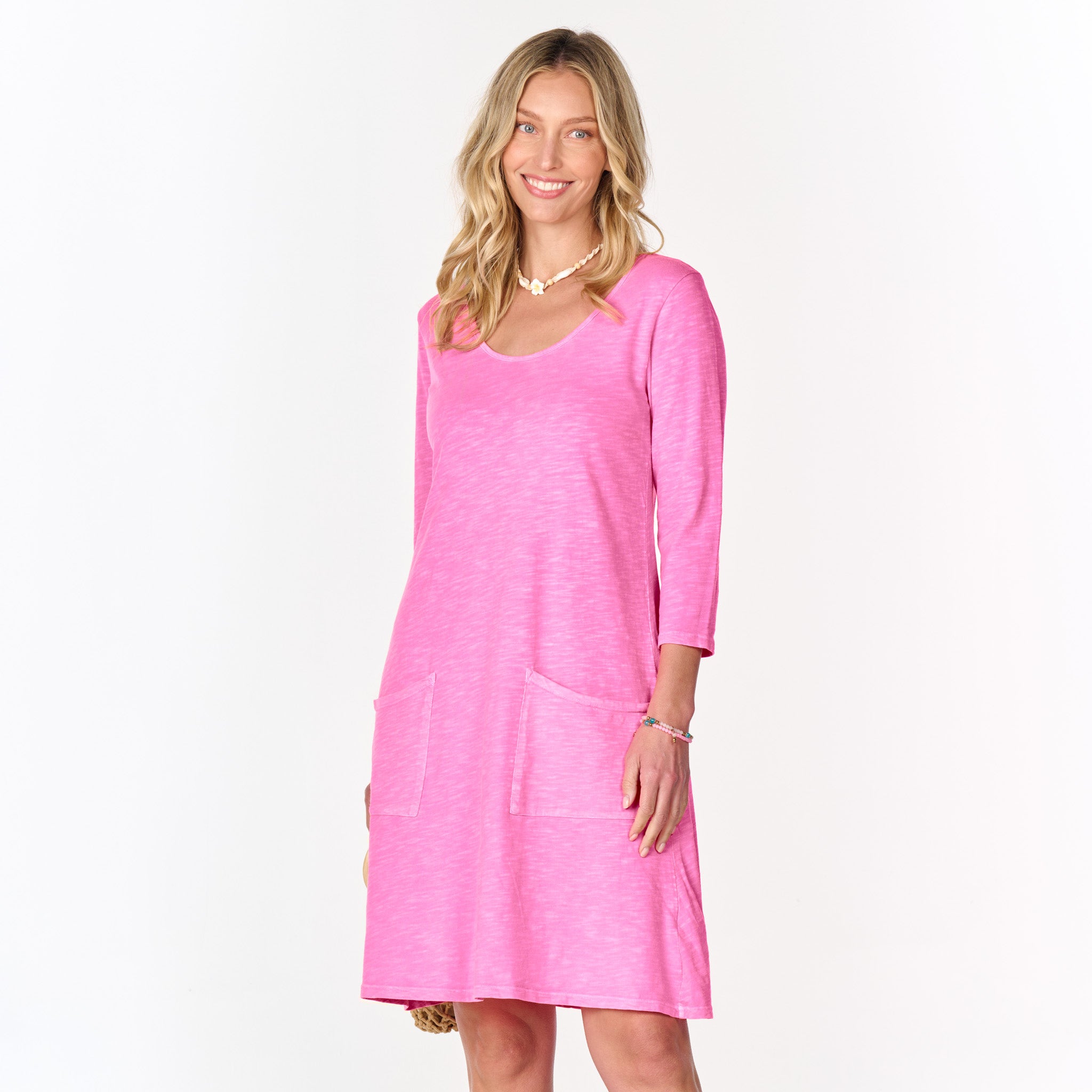 A woman with long blonde hair wears the versatile Dalia Dress by Fresh Produce Clothes in Flamingo, featuring long sleeves, front pockets, and a scoop neckline, smiling while standing against a plain white background.