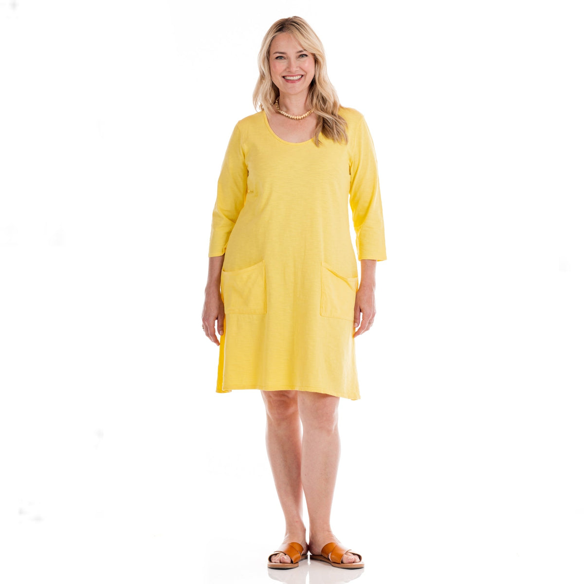 A smiling woman wears the Fresh Produce Clothes Dalia Dress | Plus Size Daffodil-Lagoon, a bright yellow knee-length cotton dress featuring three-quarter sleeves and front pockets. She stands in sandals against a white background, showcasing this stylish plus size dress proudly made in the USA.