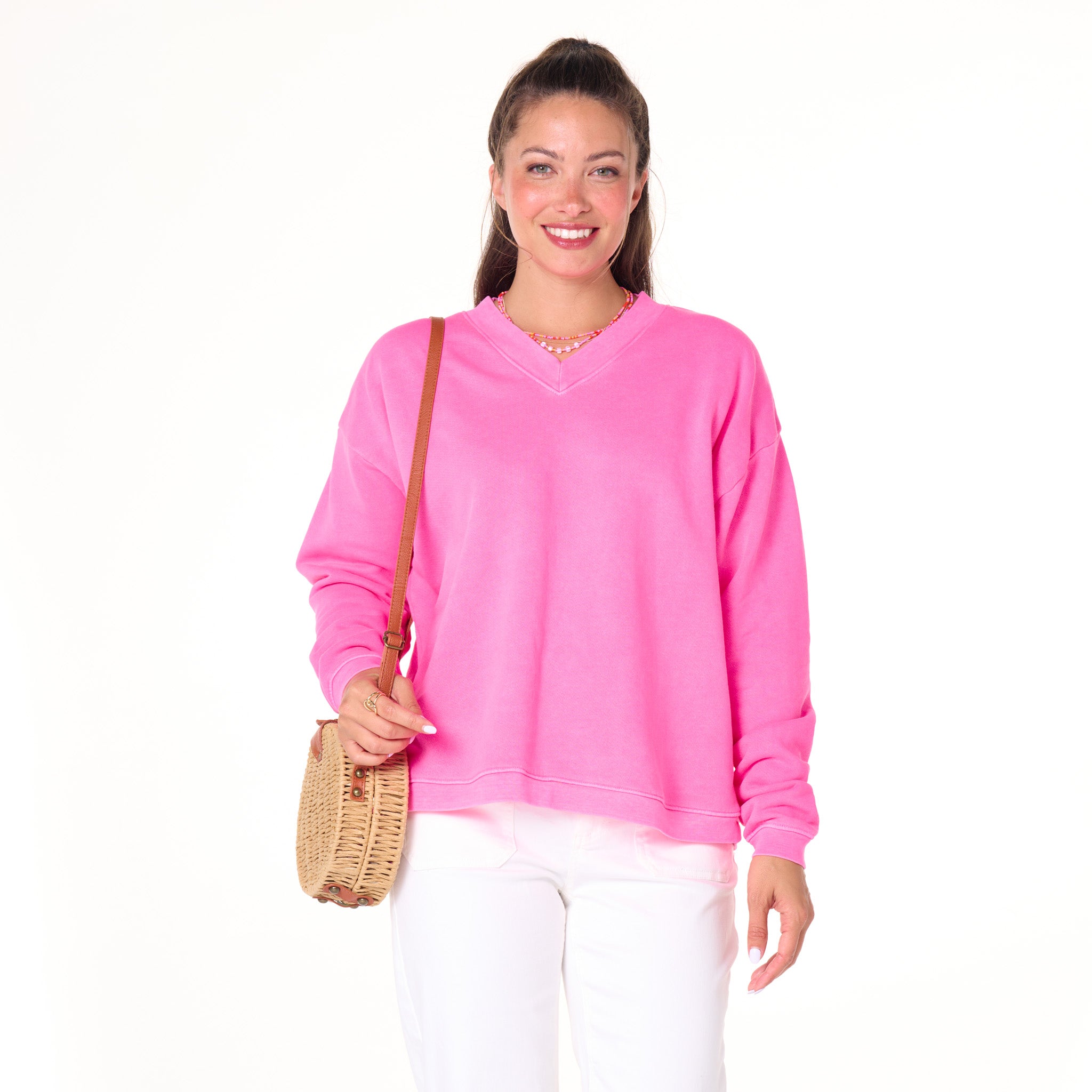 A woman wearing the Fresh Produce Clothes Crop Easy V Neck Sweatshirt in Cosmos—a bright pink shade crafted from 100% cotton fleece for a soft, lived-in feel—and white pants smiles at the camera. She carries a woven crossbody bag and has long brown hair styled half-up. The background is white and uncluttered.