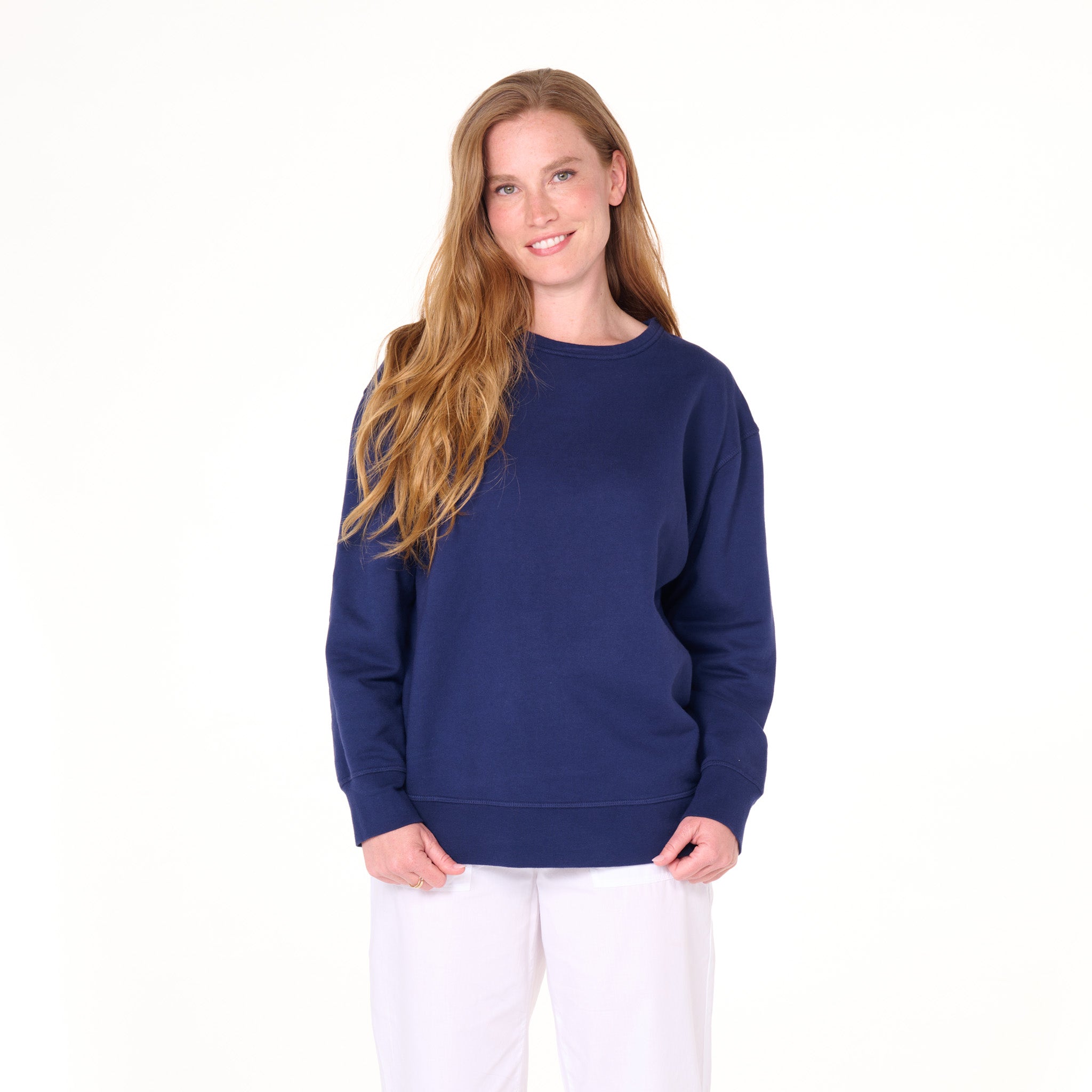 A person with long, wavy hair is smiling and wearing the Easy Sweatshirt by Fresh Produce Clothes in navy blue, paired with white pants, standing against a plain white background—the perfect look for lounging or everyday denim style.