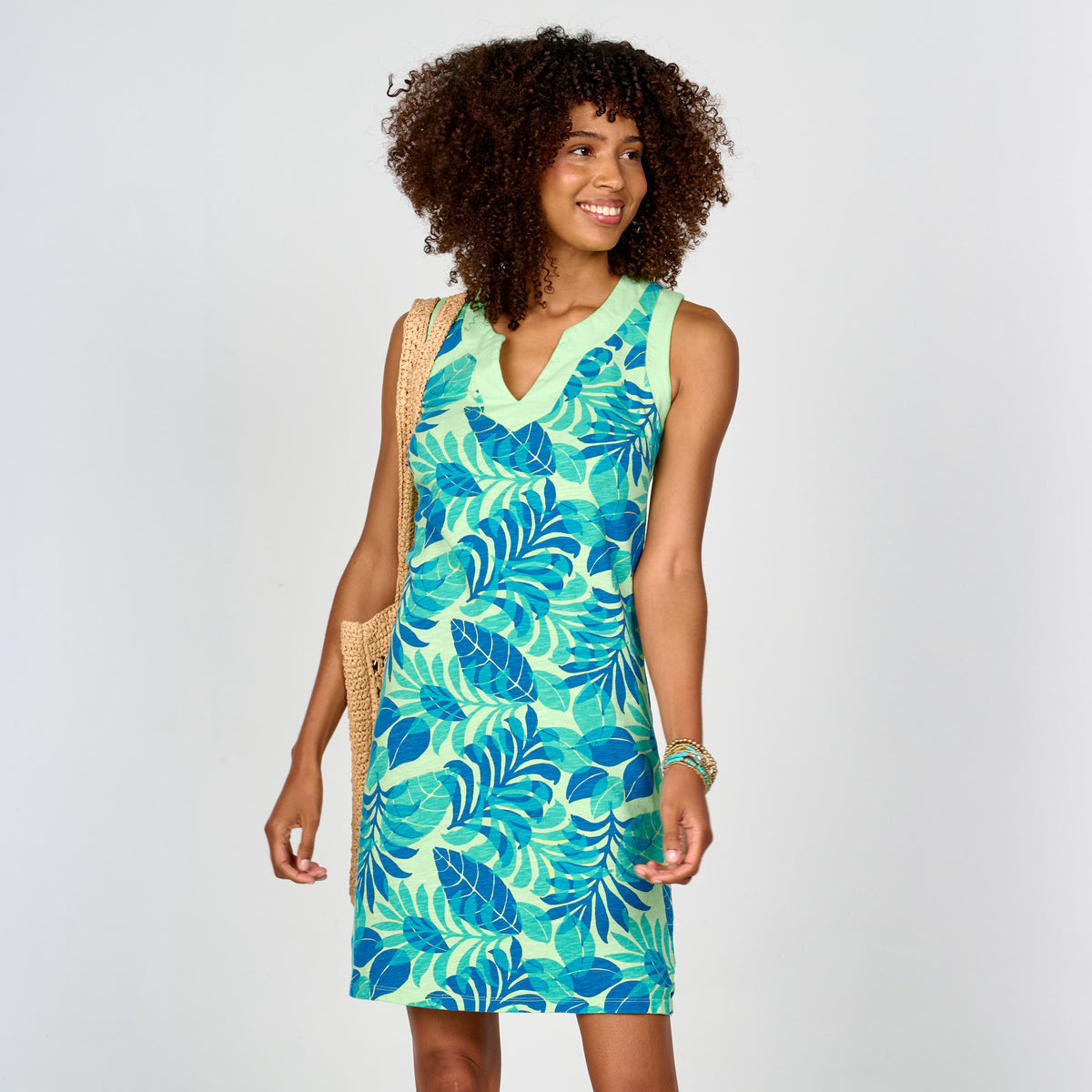 A woman with curly hair smiles and looks to the side while wearing the Fiji Palm Notcho Dress by Fresh Produce Clothes—a sleeveless, green and blue A-line dress featuring a vibrant palm print. She carries a woven bag over her shoulder against a plain white background.