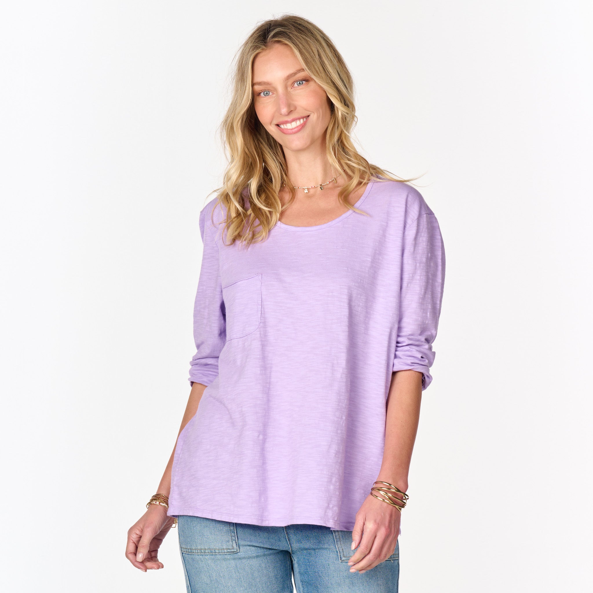 A woman with long blonde hair is smiling and standing against a plain white background, wearing the Forever Top from Fresh Produce Clothes—a loose, one-size scoop neck top in lavender—paired with blue jeans.