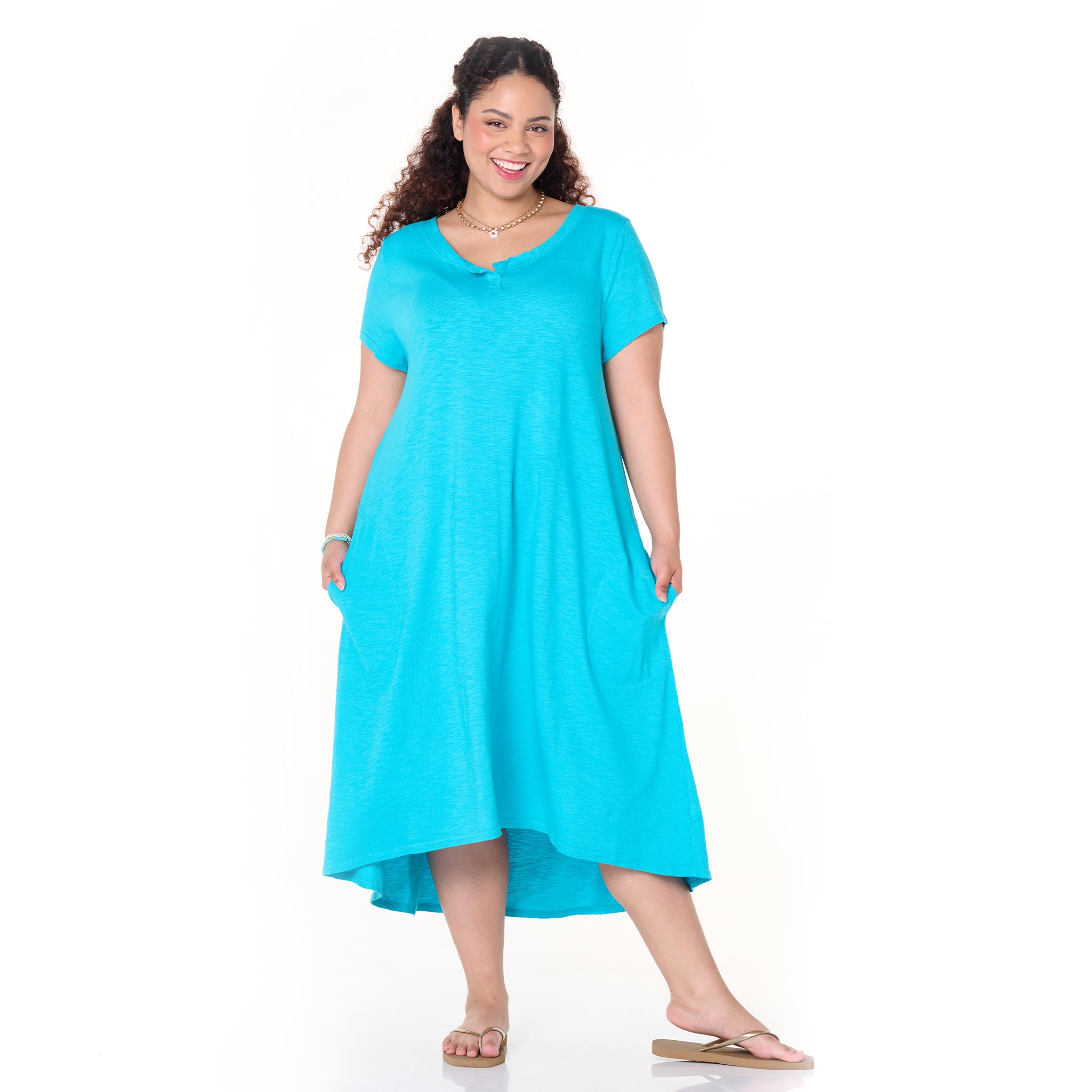 A smiling woman models the Fresh Produce Clothes Madeline Dress in Plus Size from the Clearance collection—a bright turquoise, short-sleeve midi-maxi dress with pockets and a relaxed silhouette. She accessorizes with a necklace and sandals while standing against a plain white background.