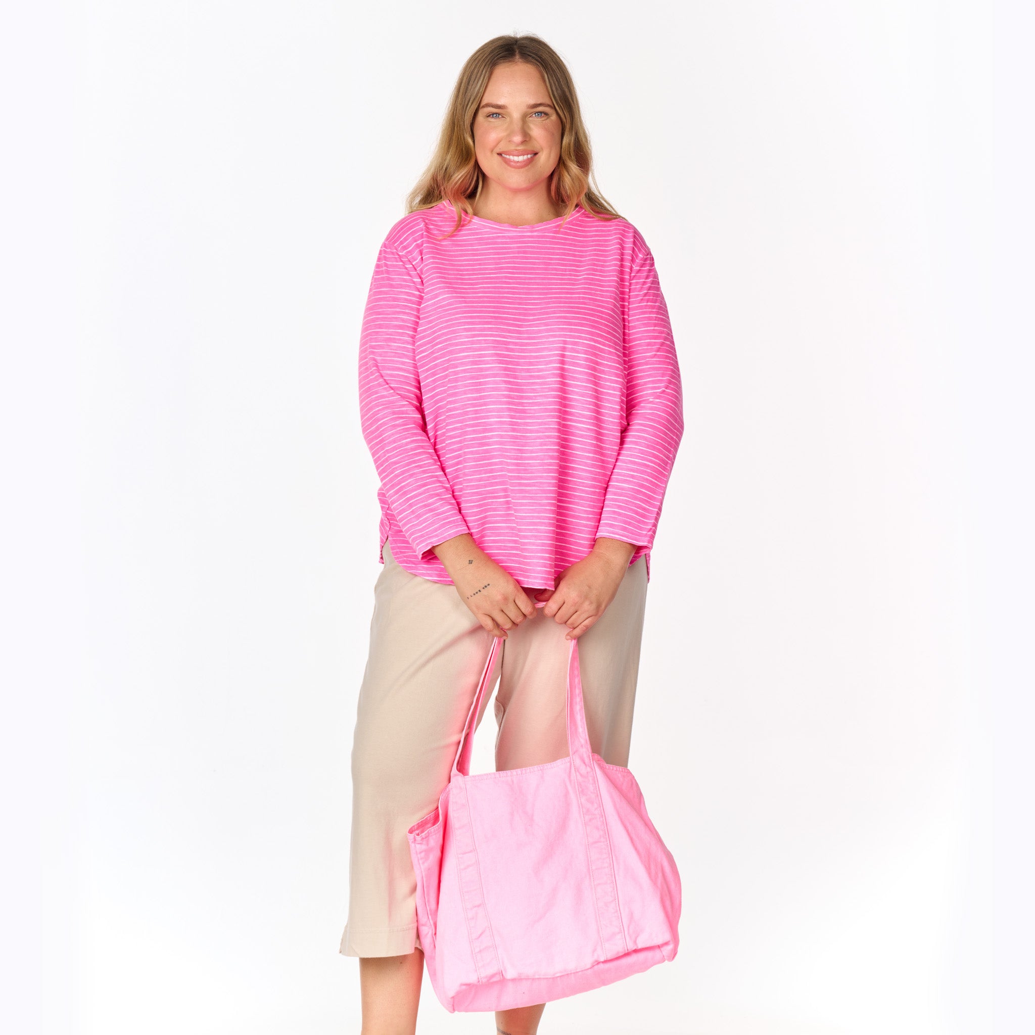 A woman smiles while wearing the Fresh Produce Clothes Ocean Stripe Catalina Top—Plus Size, in a pink and white striped print, with beige pants and holding a large light pink tote bag. She stands against a plain white background.