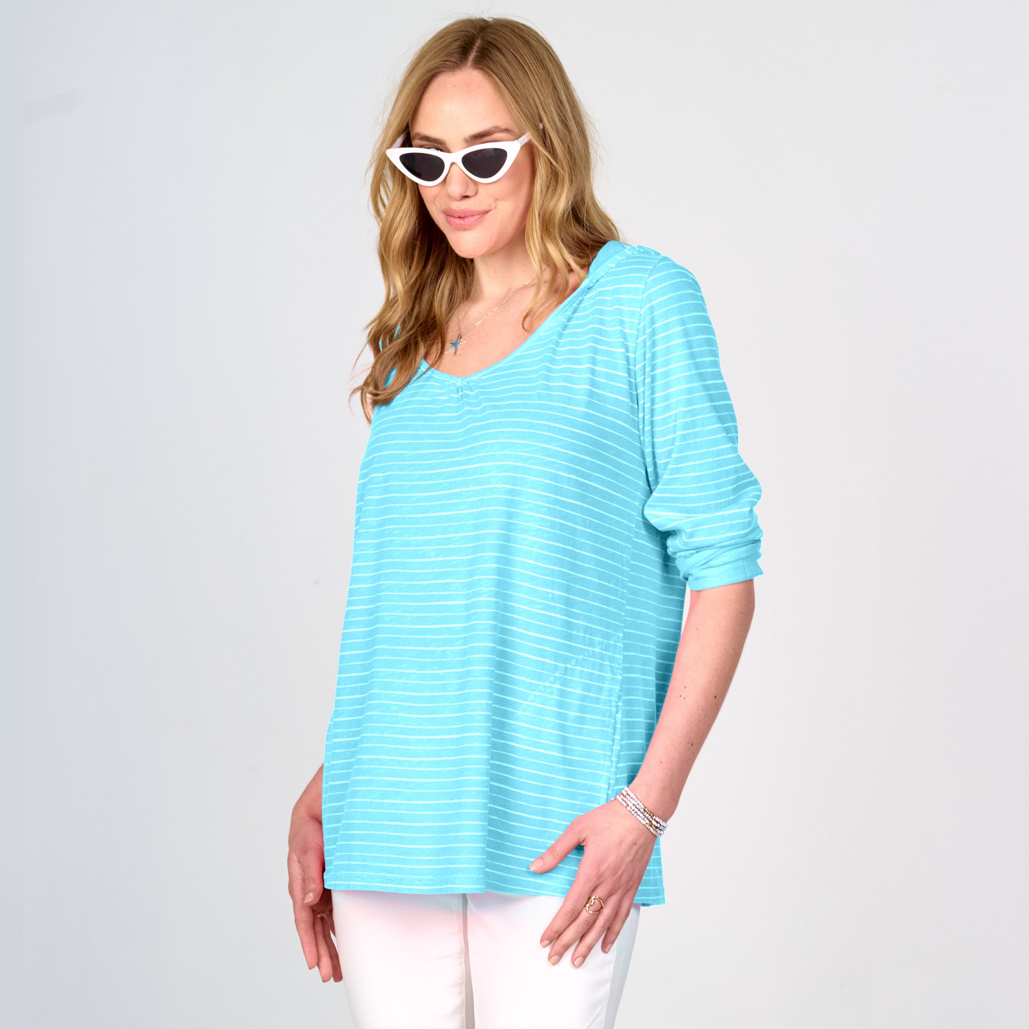 A woman wearing the Fresh Produce Clothes Ocean Stripe Hoodie | Clearance in a loose, light blue and white design made in the USA, paired with white pants, cat-eye sunglasses, and bracelets, stands against a plain light background.