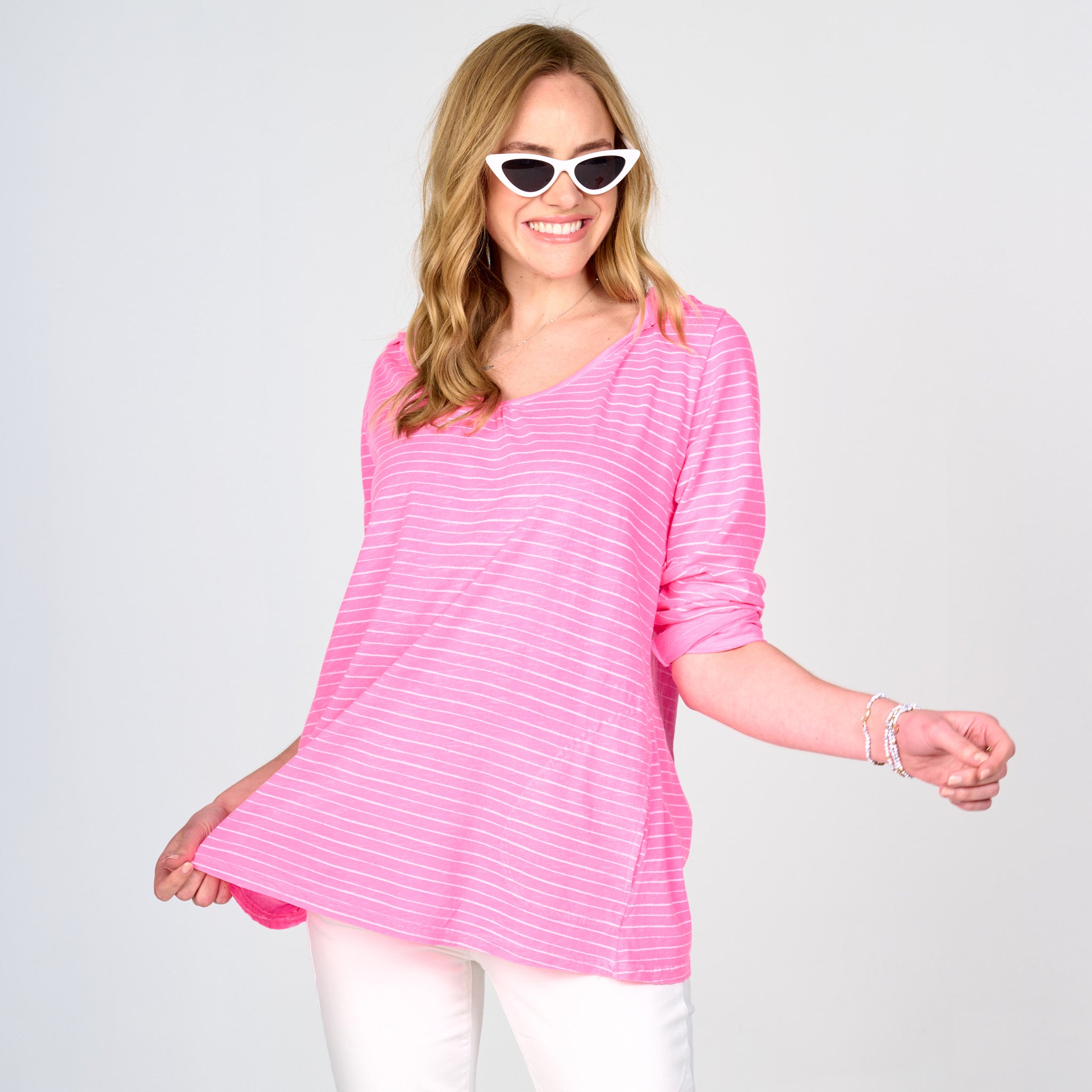 A woman with long blonde hair wears white cat-eye sunglasses, white pants, and a bracelet. She smiles and holds out one side of her Fresh Produce Clothes Ocean Stripe Hoodie—crafted from lightweight cotton in a pink and white striped design and made in the USA—against a plain light background.