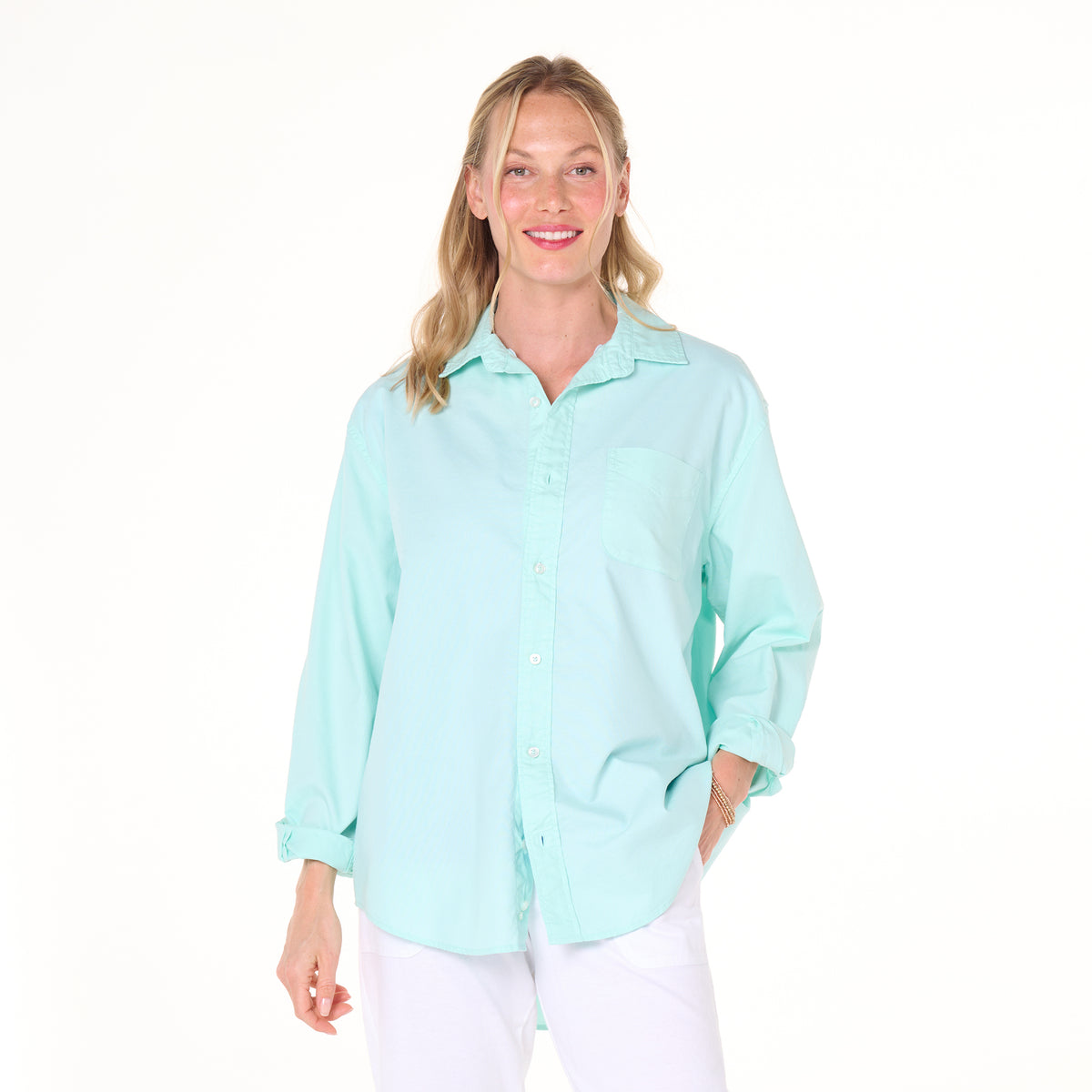 A woman with blonde hair is smiling as she stands against a plain white background, wearing the light blue Classic Oxford Shirt from Fresh Produce Clothes (Clearance) styled in an oversized fit and paired with white pants.