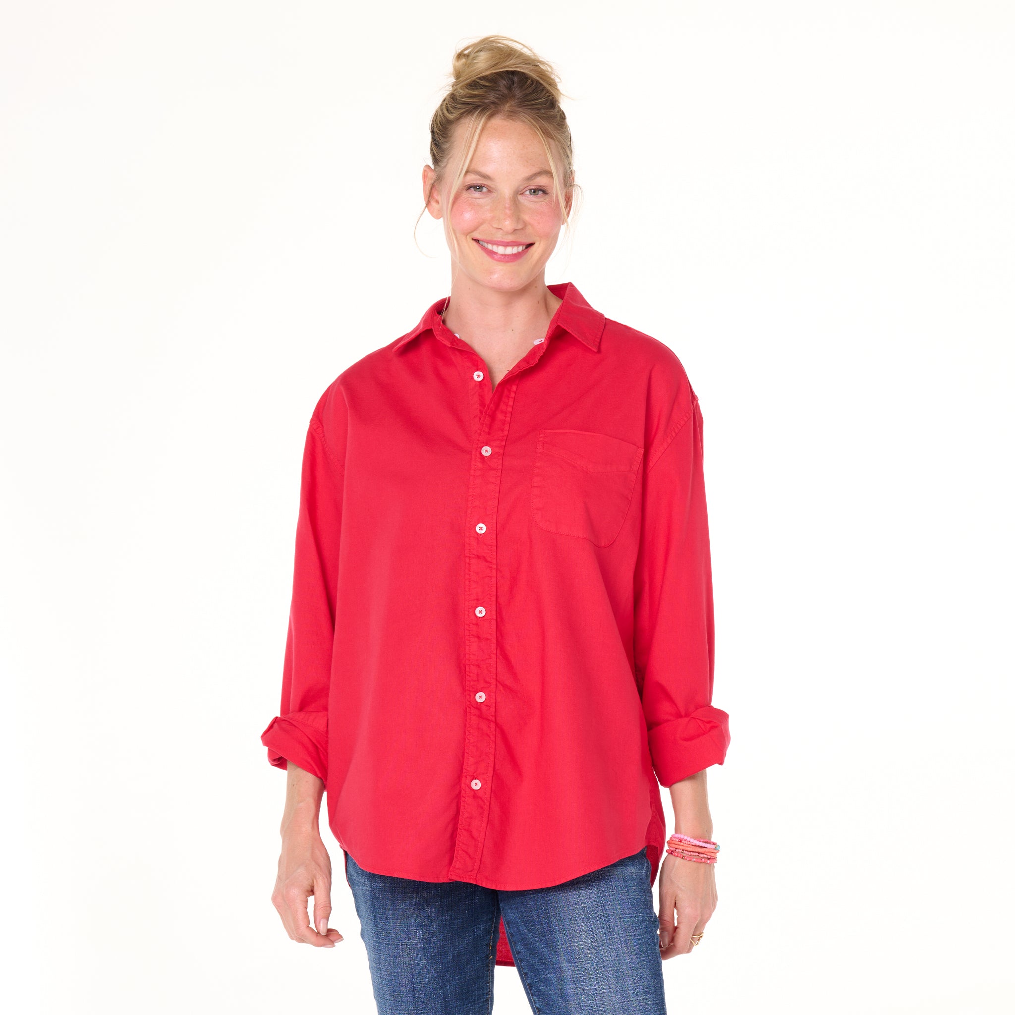 A woman with blonde hair in a bun wears the Fresh Produce Clothes Classic Oxford Shirt | Clearance in bright red, styled in a relaxed fit with rolled sleeves, paired with blue jeans and smiling against a plain white background.