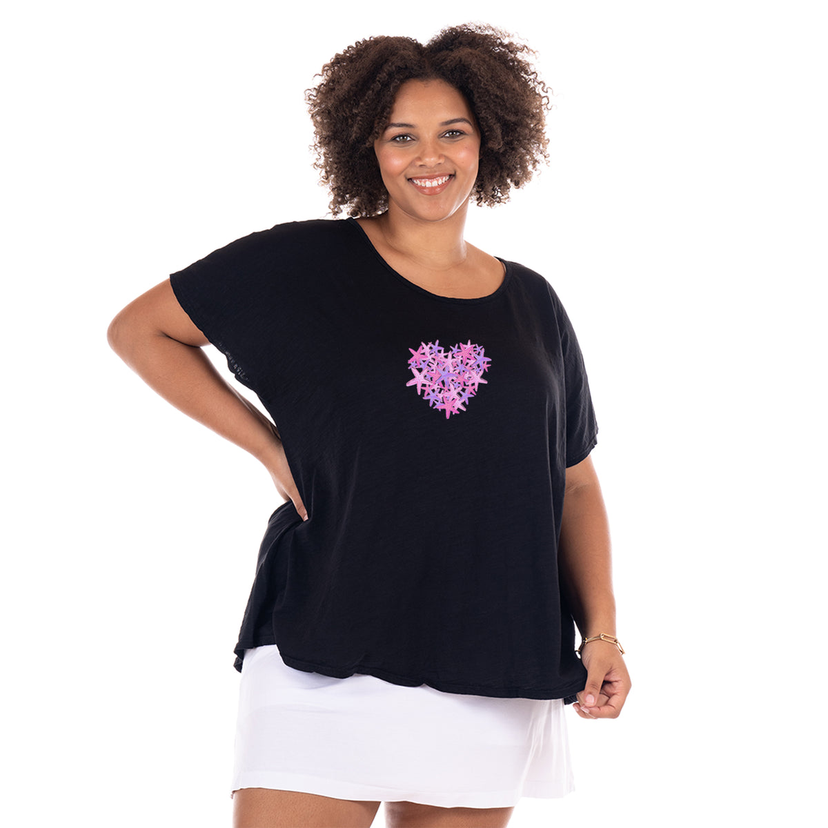 A smiling woman in the Pink Sea Heart Keepsake T by Fresh Produce Clothes—a plus size black tee featuring a pink heart design—stands with one hand on her hip, wearing a white skirt against a plain white background, making it the perfect beach gift for someone special.