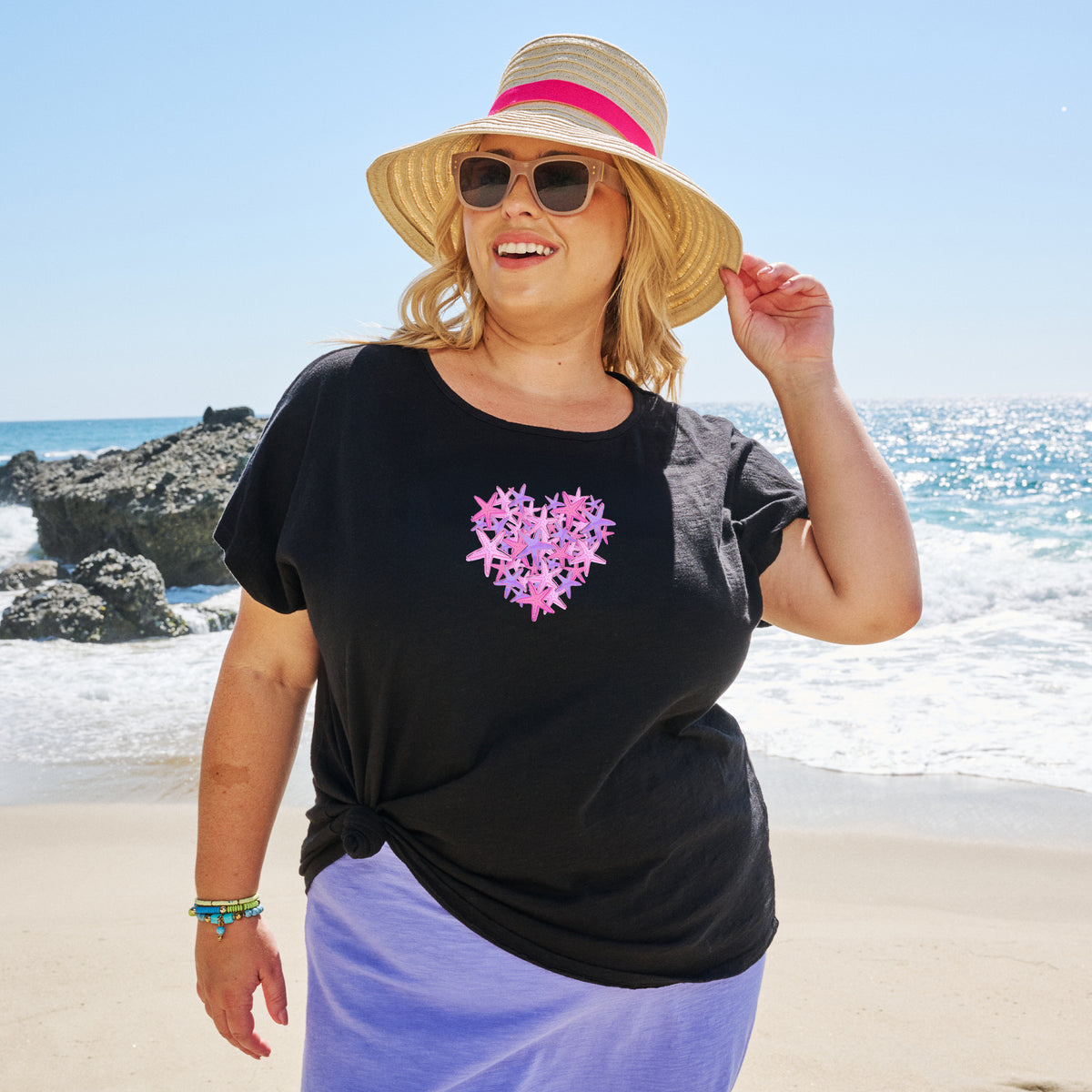 A woman in sunglasses and a straw hat smiles on a sunny beach. She wears the Pink Sea Heart Keepsake T from Fresh Produce Clothes, featuring a pink heart detail, paired with a lavender skirt. The ocean and rocks create the perfect backdrop for this lovely plus-size beach gift moment.