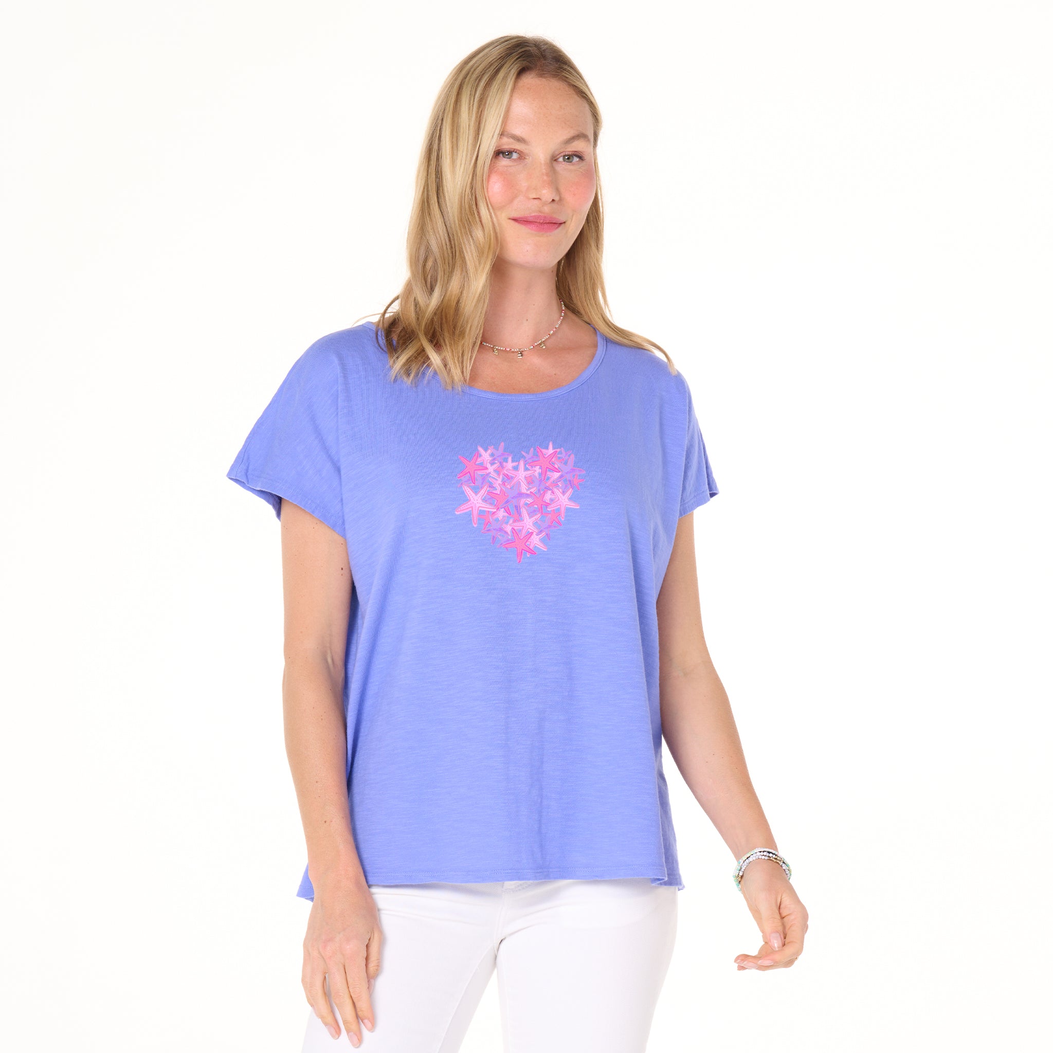 A woman wearing a Fresh Produce Clothes Pink Sea Heart Keepsake T—featuring a light purple short-sleeve design with a Pink Sea Heart graphic—paired with white pants, stands against a plain white background, smiling softly; an ideal beach gift for someone special.