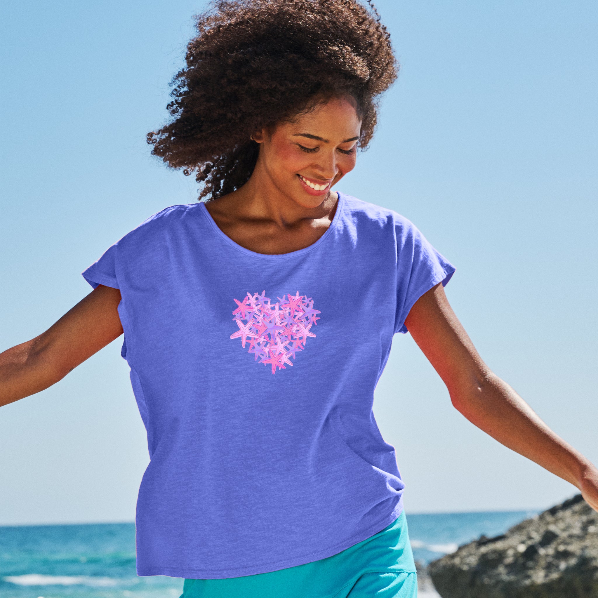 A smiling woman stands outdoors with arms outstretched, wearing the Pink Sea Heart Keepsake T by Fresh Produce Clothes; the ocean and blue sky behind her create a perfect backdrop for this thoughtful beach gift.