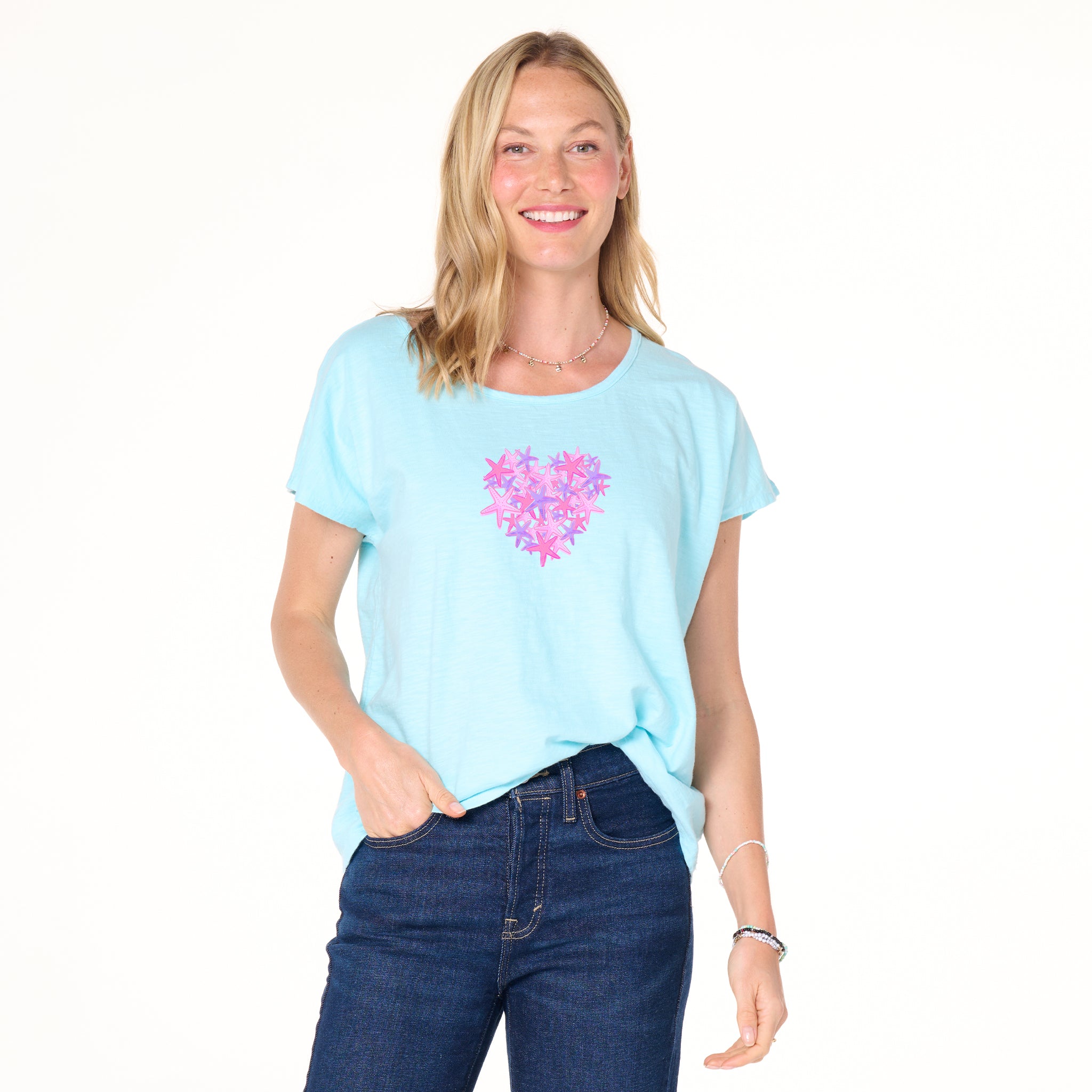 Smiling woman in a light blue Pink Sea Heart Keepsake T by Fresh Produce Clothes, featuring a pink and purple heart design, paired with dark blue jeans, standing against a white background—the perfect beach gift.