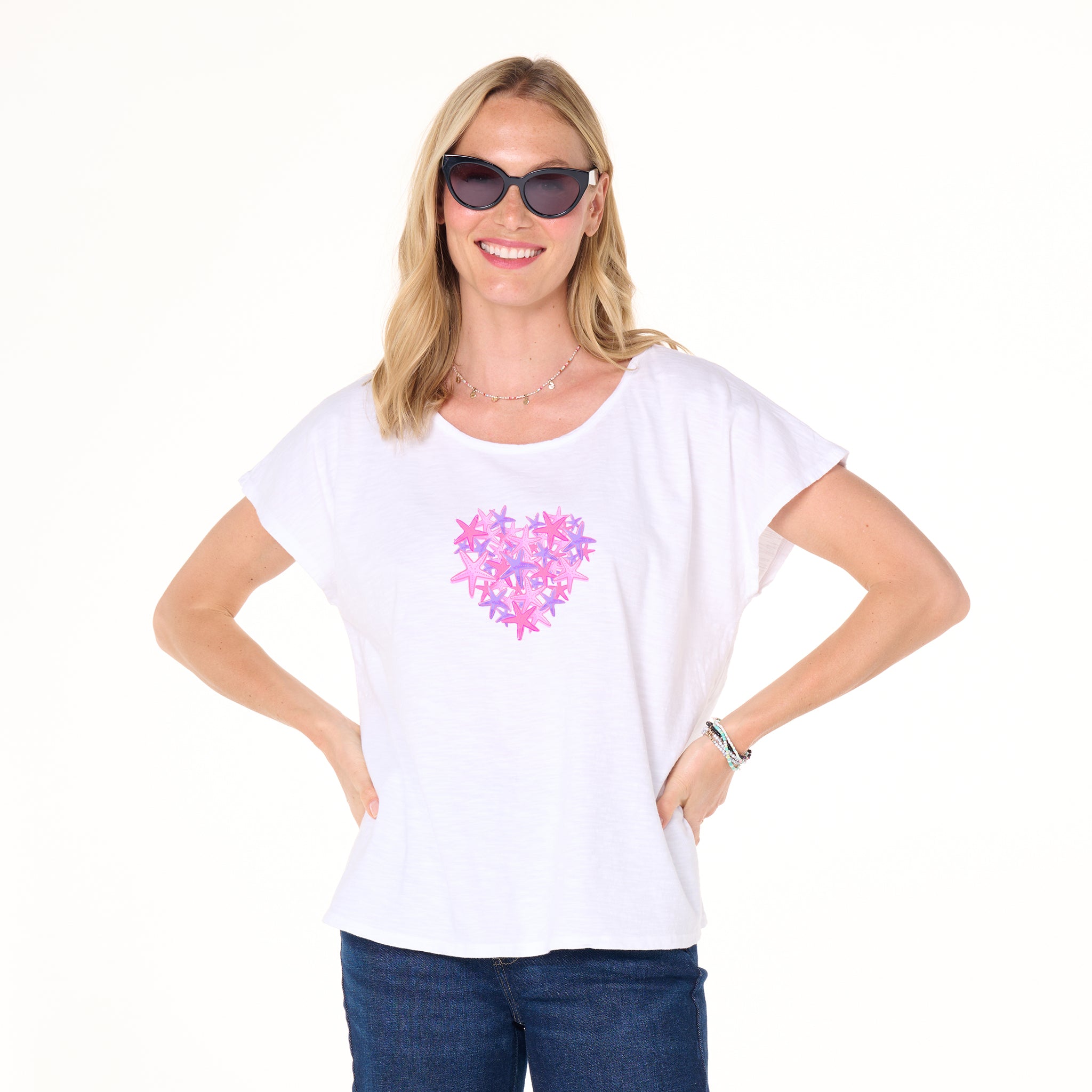 A smiling woman in sunglasses and a white t-shirt adorned with the Pink Sea Heart floral design stands with hands on her hips against a white background. The Pink Sea Heart Keepsake T by Fresh Produce Clothes makes an ideal beach gift for someone special.