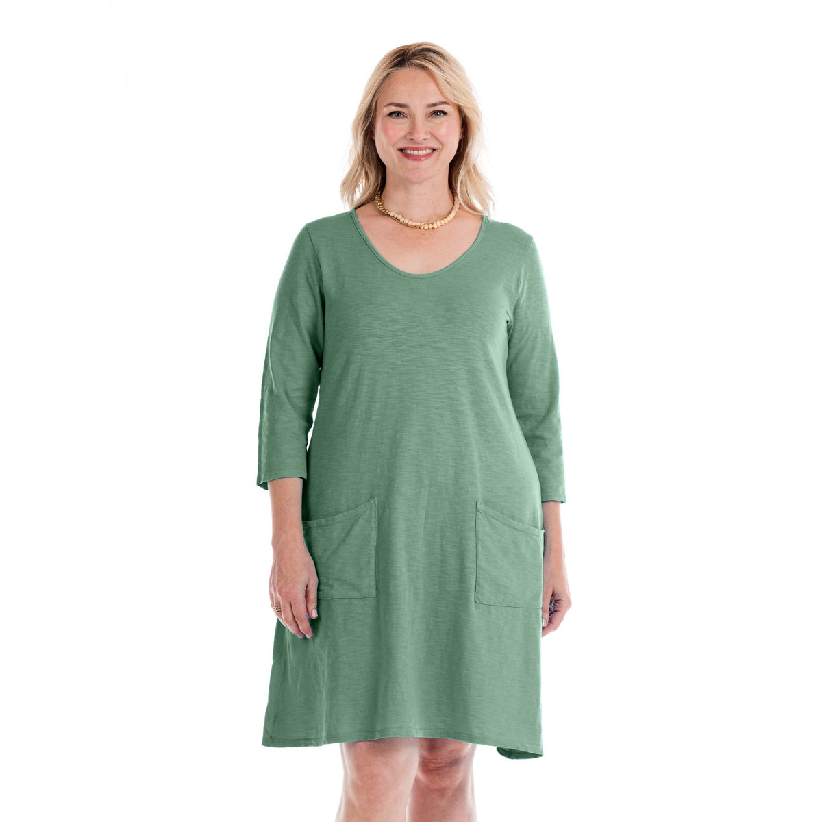A woman wearing the Dalia Dress | Plus Size Daffodil-Lagoon by Fresh Produce Clothes—a light green, knee-length cotton dress with three-quarter sleeves and front pockets—stands smiling against a plain white background. This stylish plus size dress is proudly made in the USA.