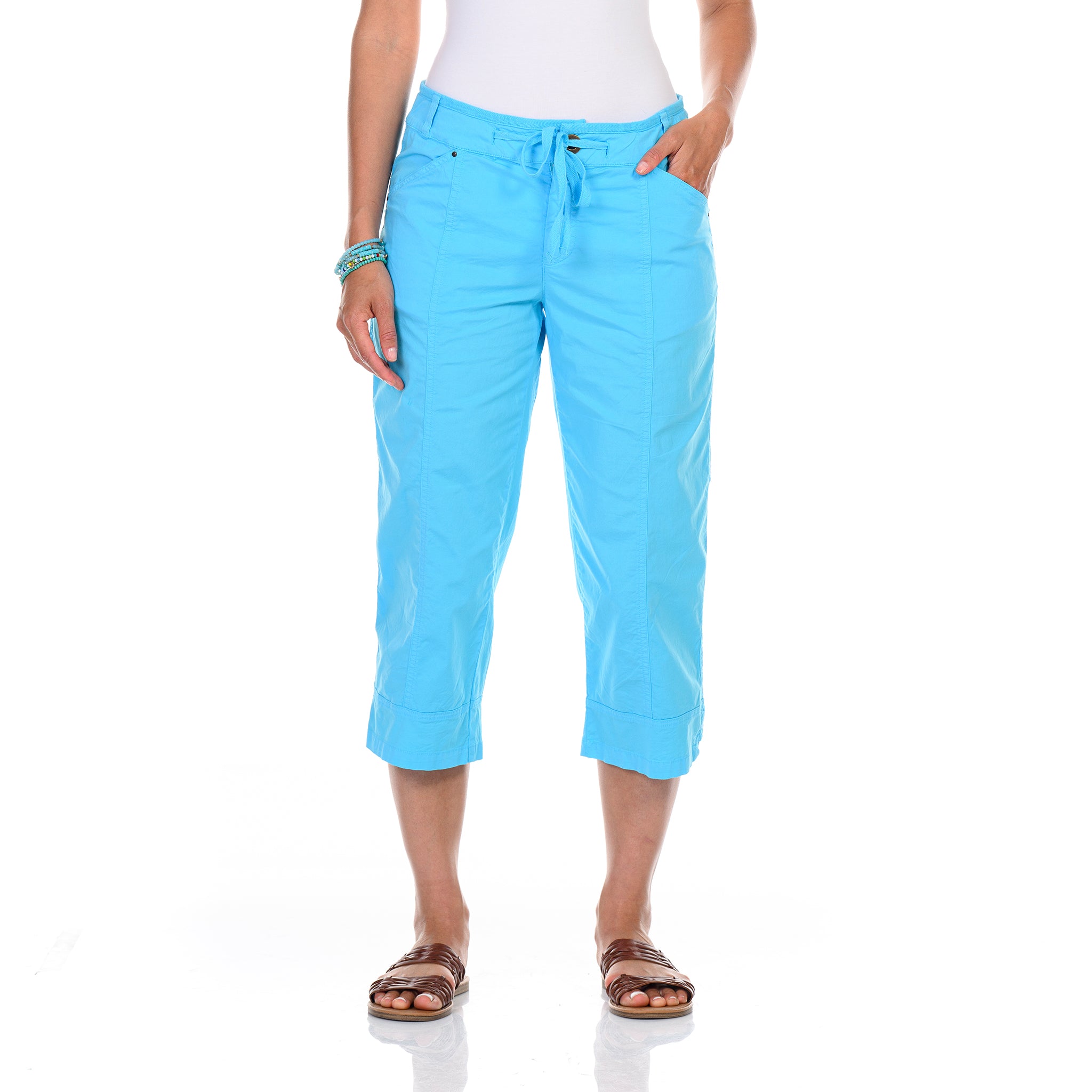 A person stands with hands in pockets against a white background, wearing Fresh Produce Clothes’ Safari Capris in Blue Fin—a bright blue capri made from stretch broadcloth with a comfortable drawstring waist—paired with a white sleeveless top, brown sandals, and bracelets.