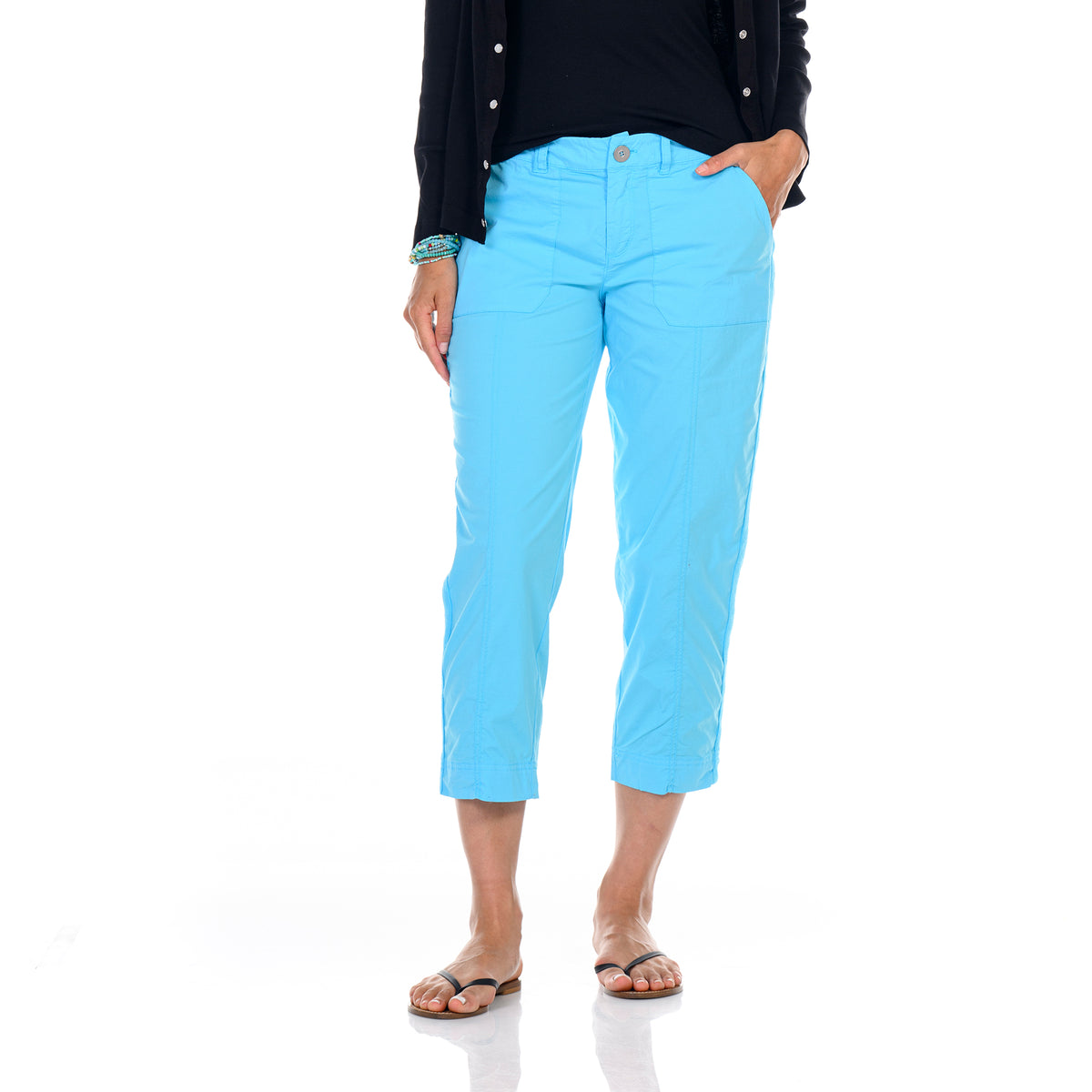 A person wearing Fresh Produce Clothes Safari Pocket Capris in Blue Fin, featuring subtle embroidery stitch details, pairs them with a black top, black cardigan, and sandals while standing with one hand in a pocket against a white background.