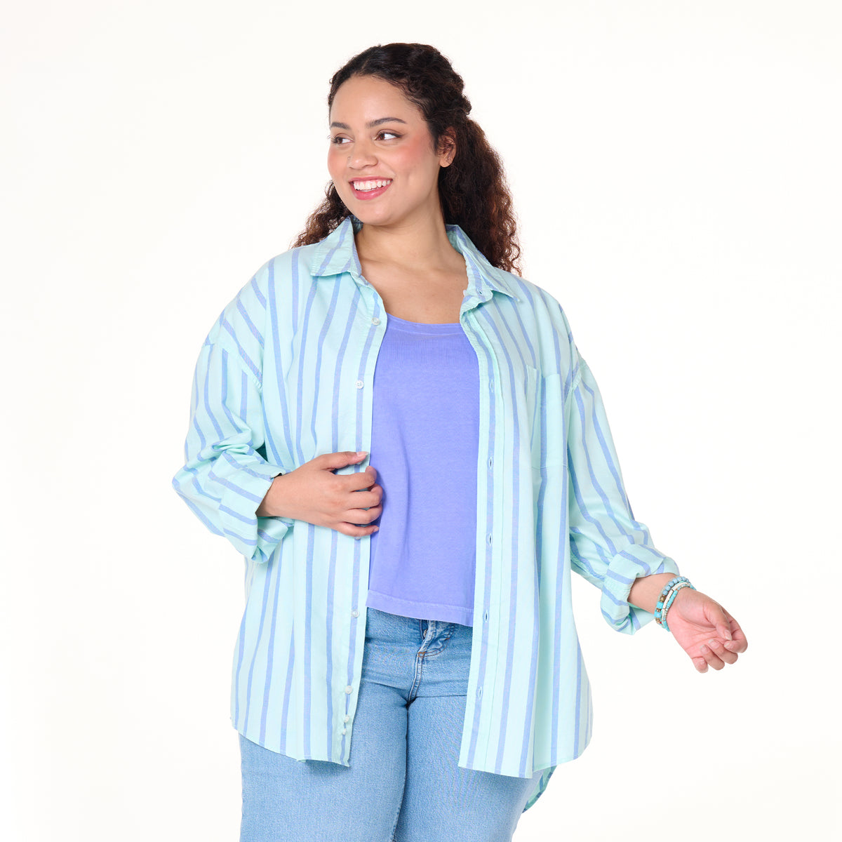 A woman with curly hair smiles as she poses against a white background, wearing the Fresh Produce Clothes Sailaway Oxford Shirt in plus size—a relaxed fit blue and white striped button-down—layered over a purple top and paired with light blue jeans.