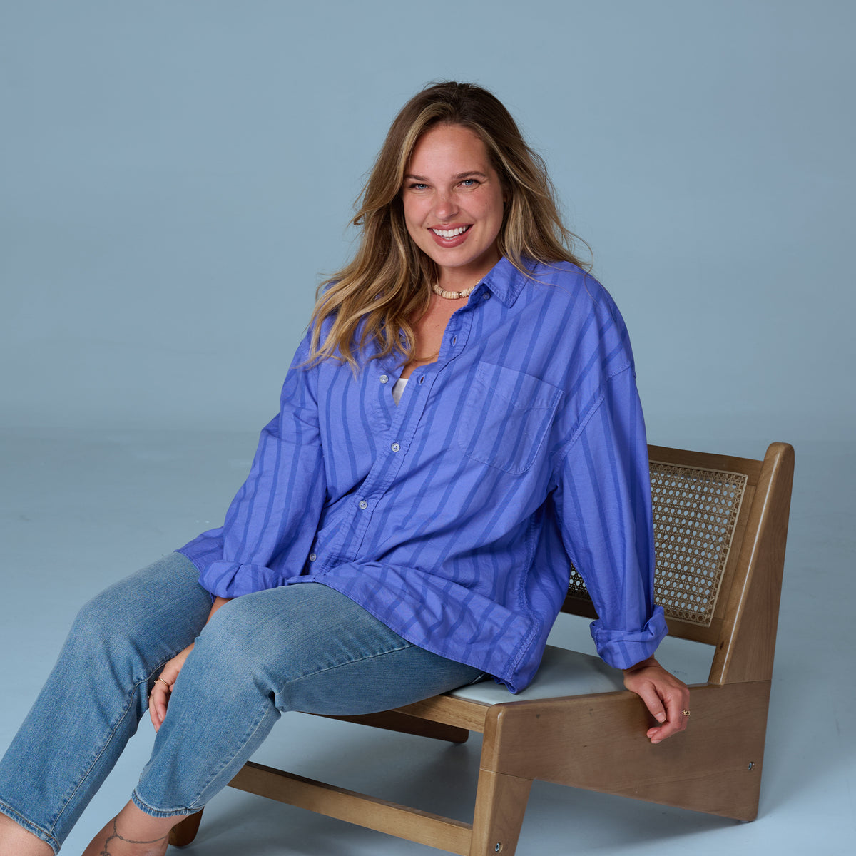 A woman with long blonde hair, wearing the Sailaway Oxford Shirt | Plus Size in blue stripes from Fresh Produce Clothes and relaxed fit jeans, sits sideways on a wooden chair, smiling at the camera against a plain light blue background.