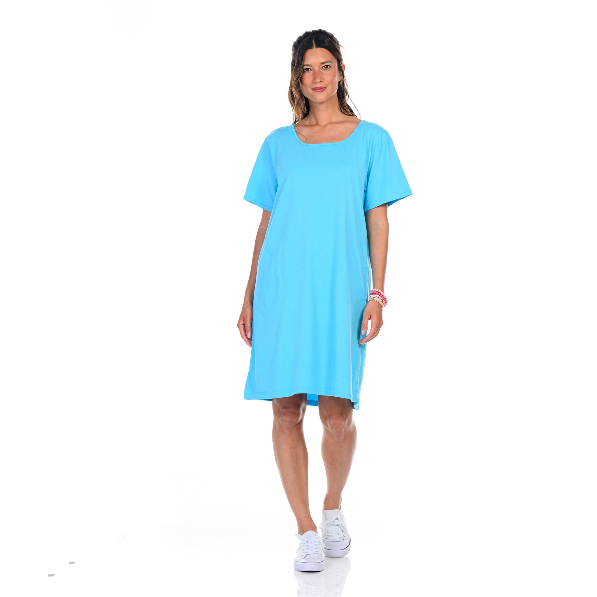 A woman stands against a white background, wearing the Fresh Produce Clothes Sanibel Dress in Blue Fin—a loose-fitting, short-sleeved t-shirt dress crafted from soft cotton jersey. Her long hair is styled in a ponytail and she smiles gently, with one hand by her side and the other relaxed. The dress features convenient pockets, and she completes her look with crisp white sneakers.