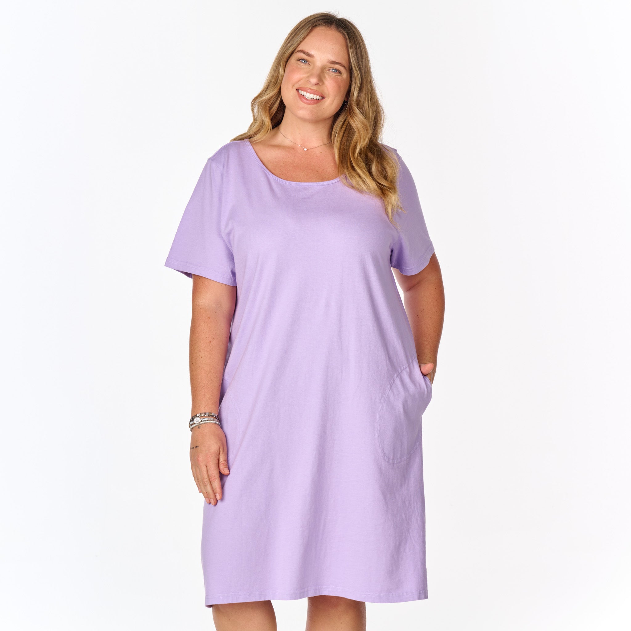 A woman with long, light brown hair smiles while wearing the Sanibel Dress by Fresh Produce Clothes—a plus size, knee-length, short-sleeve lavender cotton jersey dress—posing with her hand in one pocket against a plain white background.