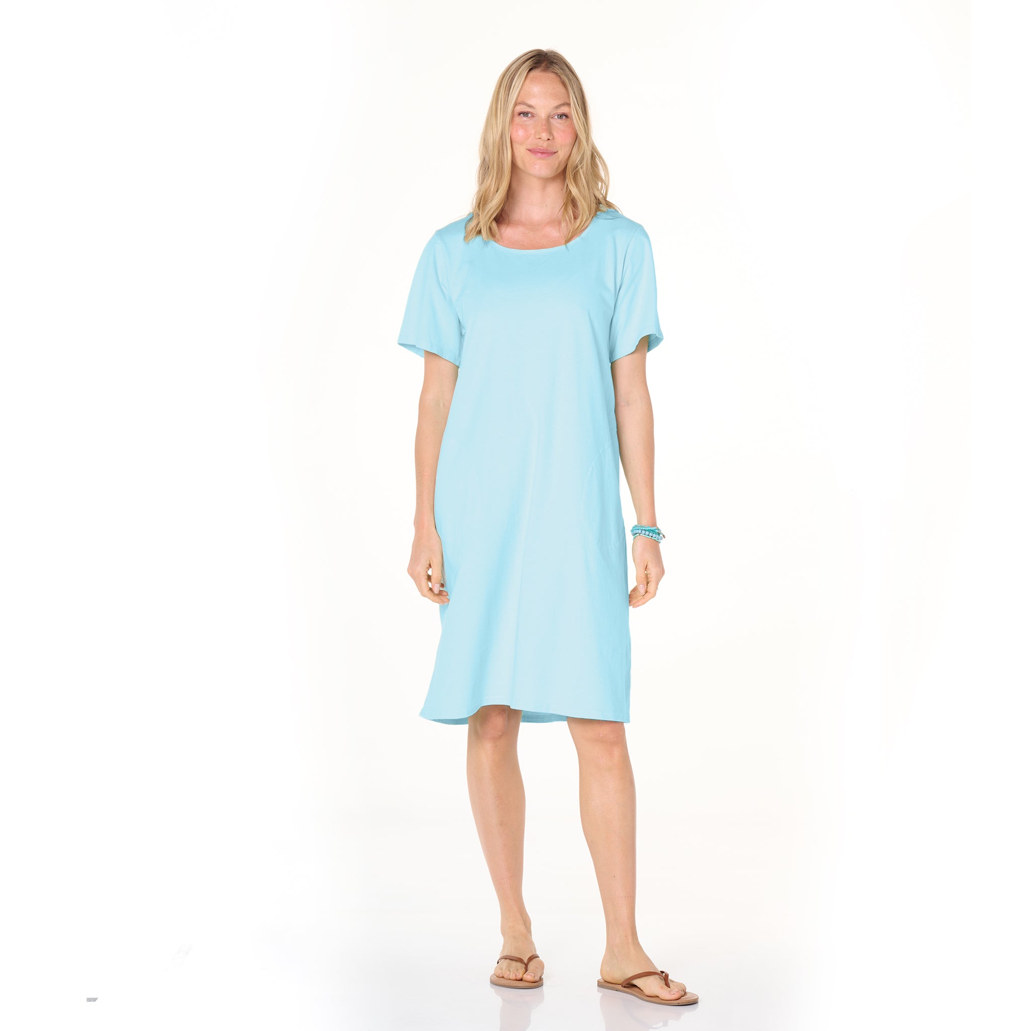 A woman stands facing forward, wearing the Fresh Produce Clothes Sanibel Dress in light blue, styled as an oversized t-shirt dress with short sleeves and paired with sandals—perfect for all-day wear—against a light background.