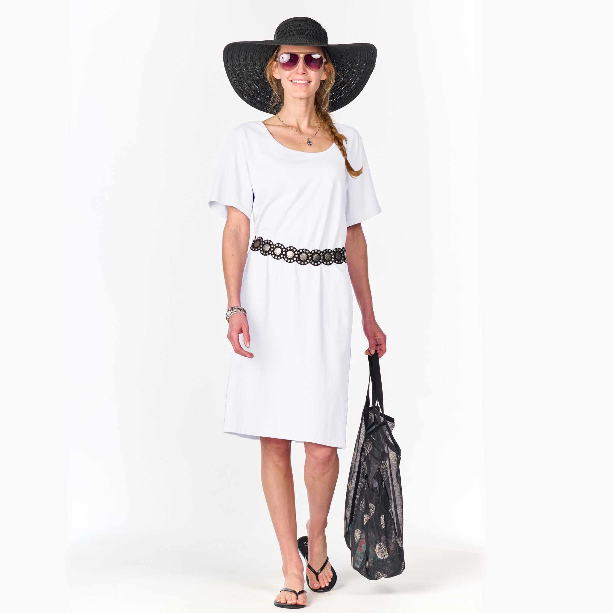 Wearing the Fresh Produce Clothes Sanibel Dress in white, paired with a black sunhat, sunglasses, a black bag, and flip-flops, she smiles against a plain white background—an effortlessly stylish look perfect for all-day wear.