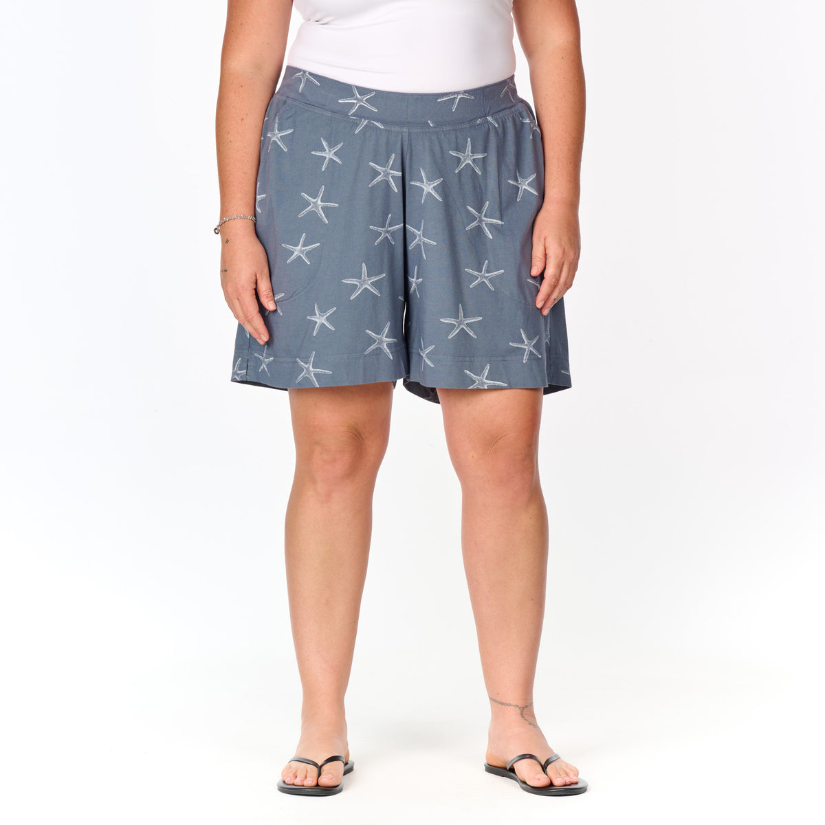 A person wearing a white top and the Fresh Produce Clothes Sea Star 7″ Key Largo Shorts in plus size, featuring a starfish pattern, stands on a white background with black flip-flops.